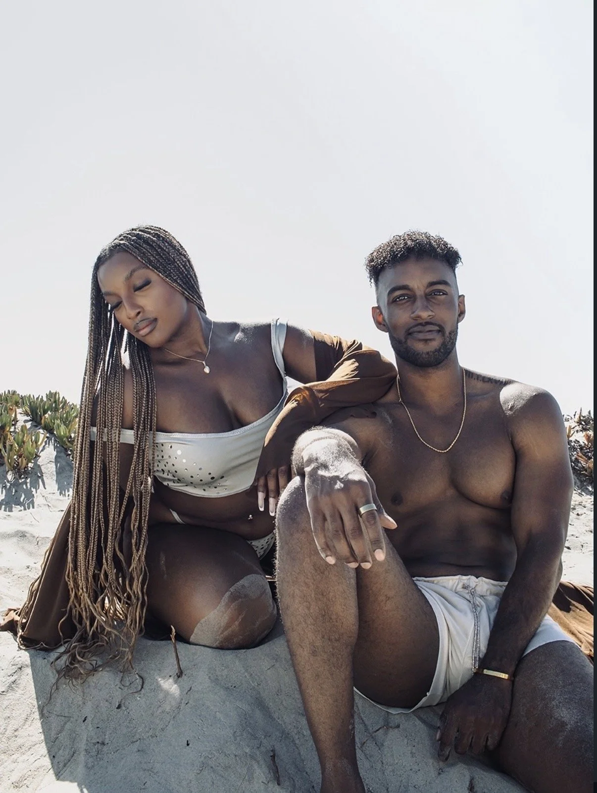 A man and a woman sitting on the beach, surrounded by sand and some green plants, with clear sky in the background. The woman has long braided hair and is wearing a white top, while the man is shirtless, wearing white shorts and sitting with one arm resting on his knee.