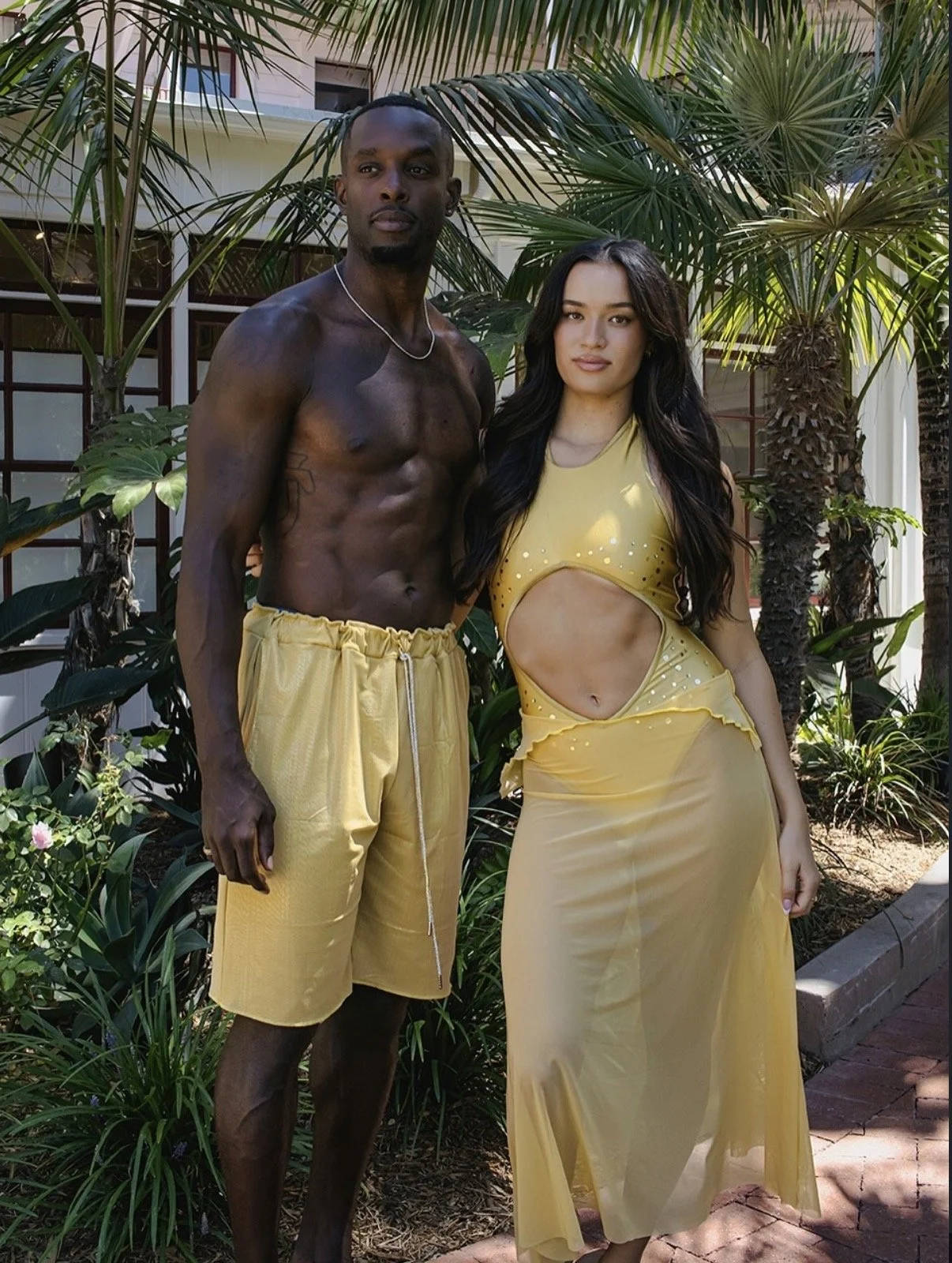 A shirtless man wearing yellow shorts standing next to a woman in a yellow dress with a cutout design, posing outdoors among palm trees and plants.