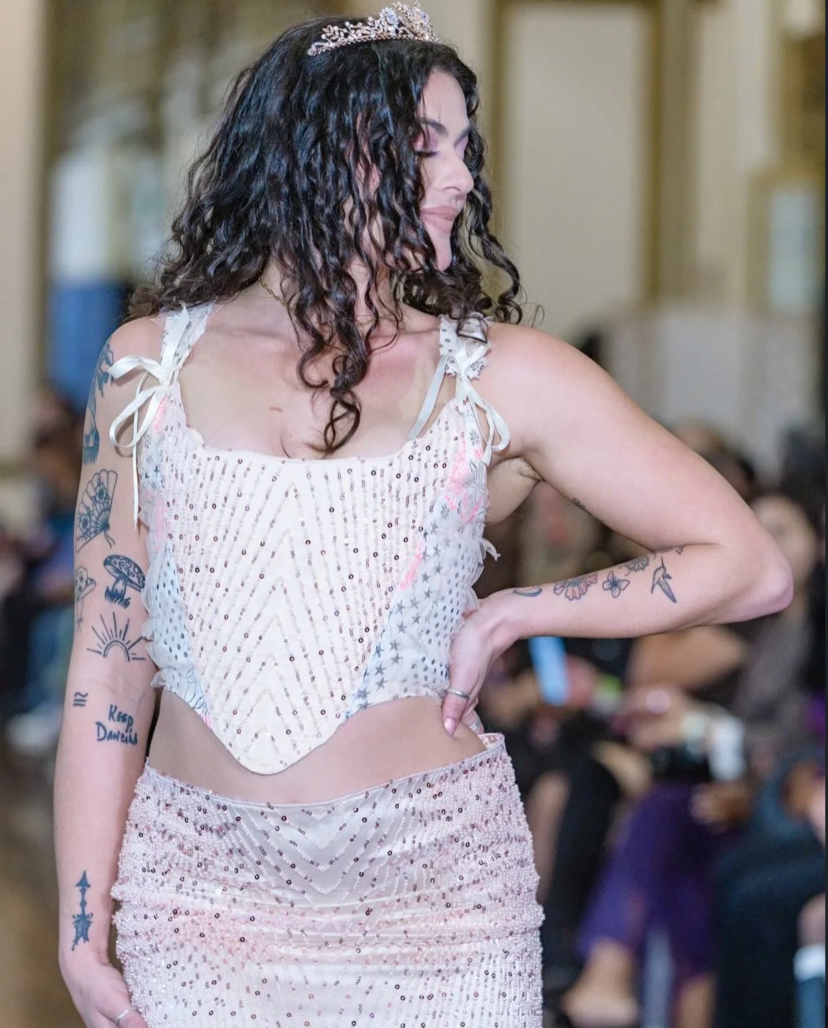 Woman with dark curly hair wearing a white, sequined dress and tiara, standing in front of a blurred crowd.