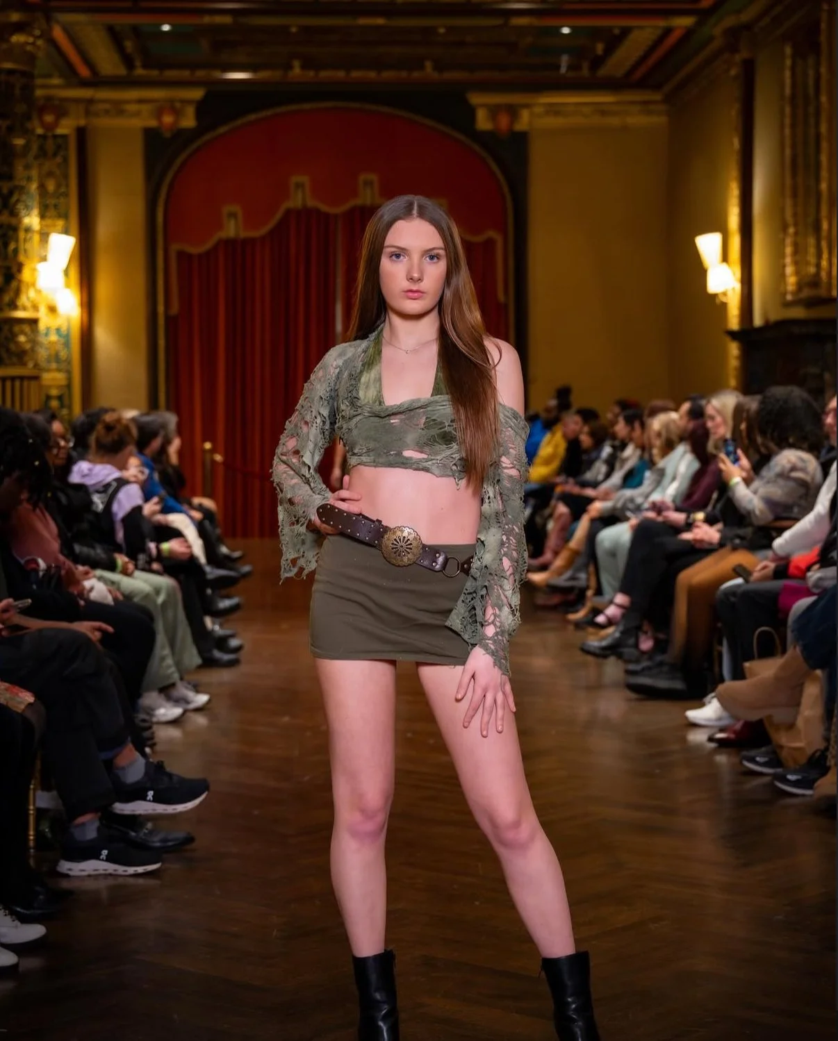 A young woman confidently walking on a fashion runway in a studio setting with an audience on either side, wearing a green tattered crop top, a matching mini skirt, black boots, and a belt with a large decorative buckle.
