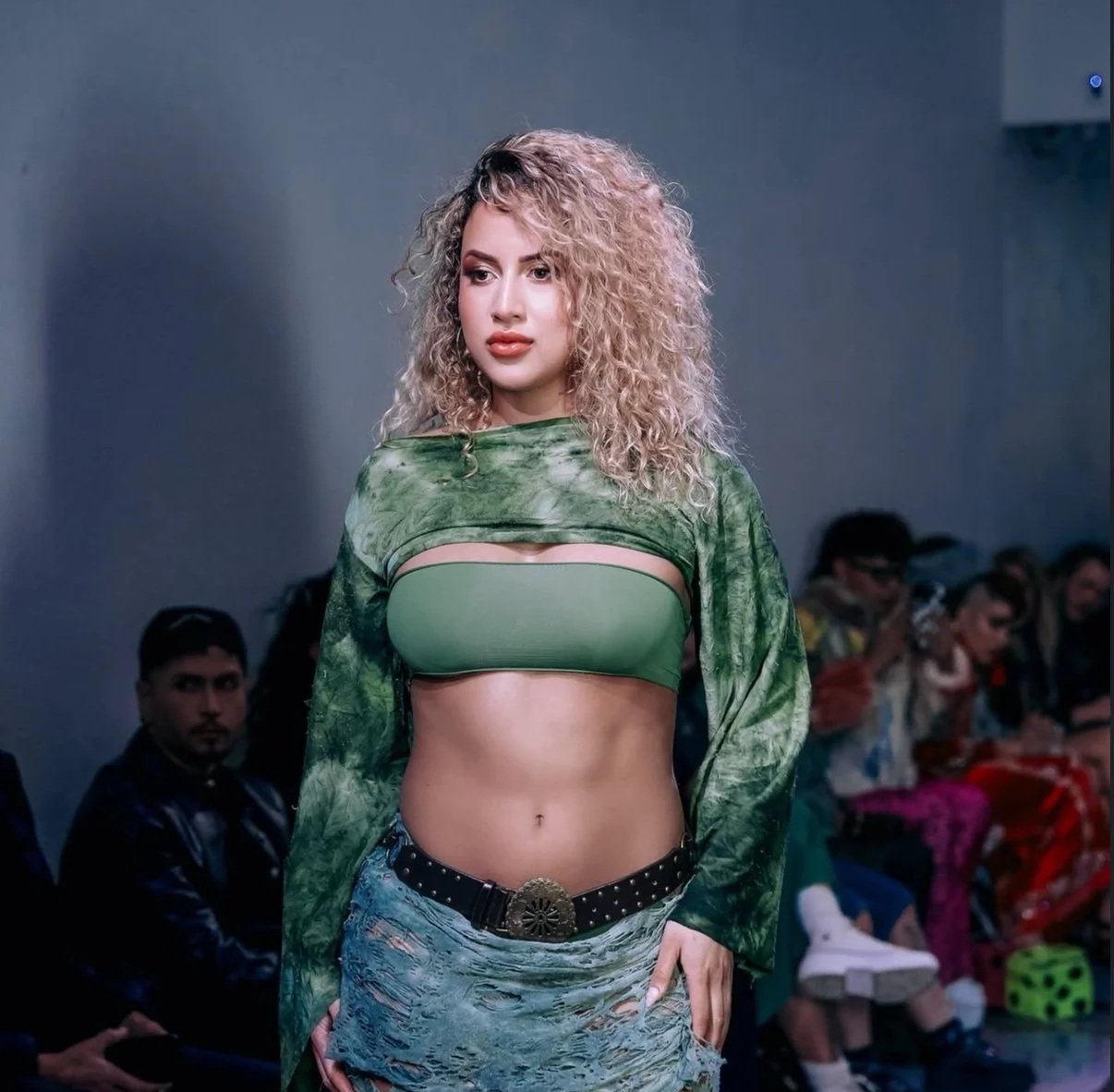 A young woman with curly blonde hair walking on a runway in a green and tie-dye crop top, distressed denim shorts, and a belt with a large circular buckle, during a fashion show with people seated in the background.