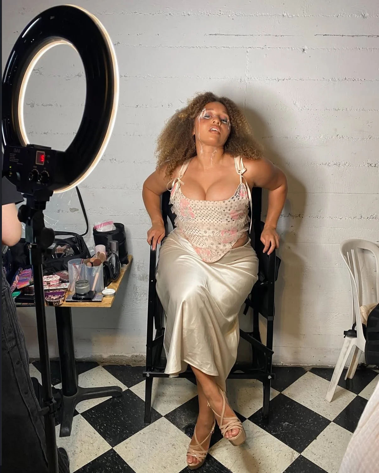 A woman with curly hair and a floral top sitting on a chair during a photoshoot, with makeup and beauty products on a table nearby and a ring light illuminating the scene.