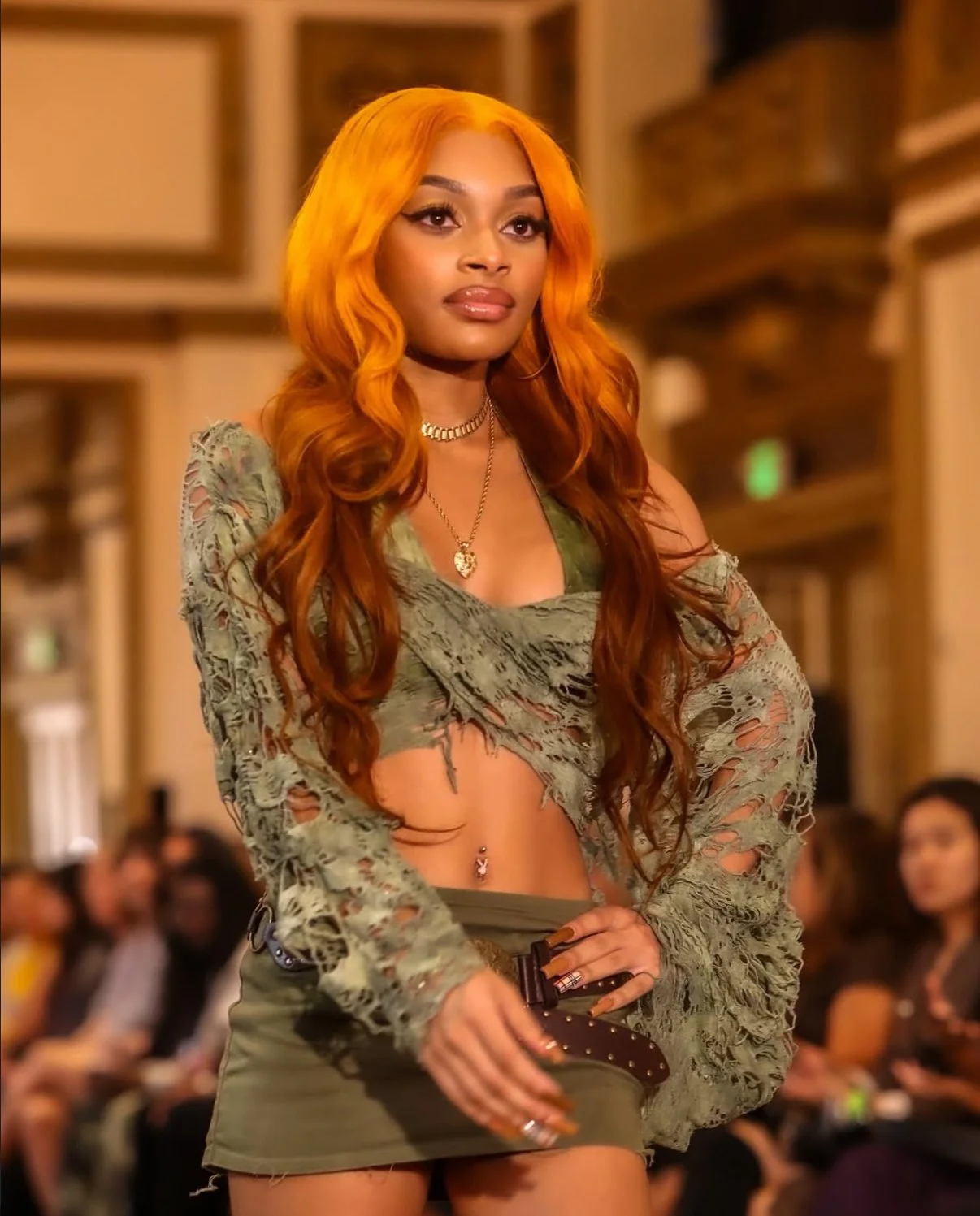A young woman with long, wavy orange hair walking on a runway, wearing a distressed green crop top, a green mini skirt, a belt, jewelry, and holding a small purse, with an audience in the background.