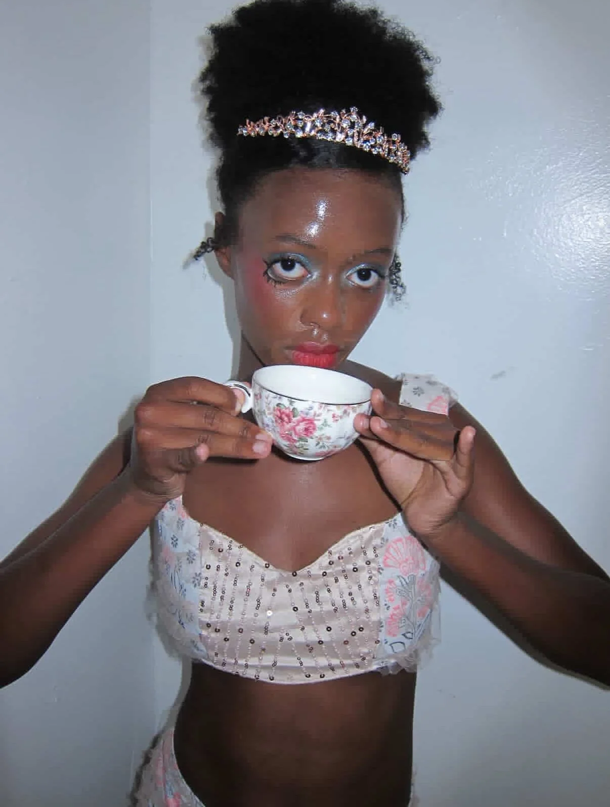 Young woman with makeup and a tiara holding a floral teacup, looking directly at the camera.