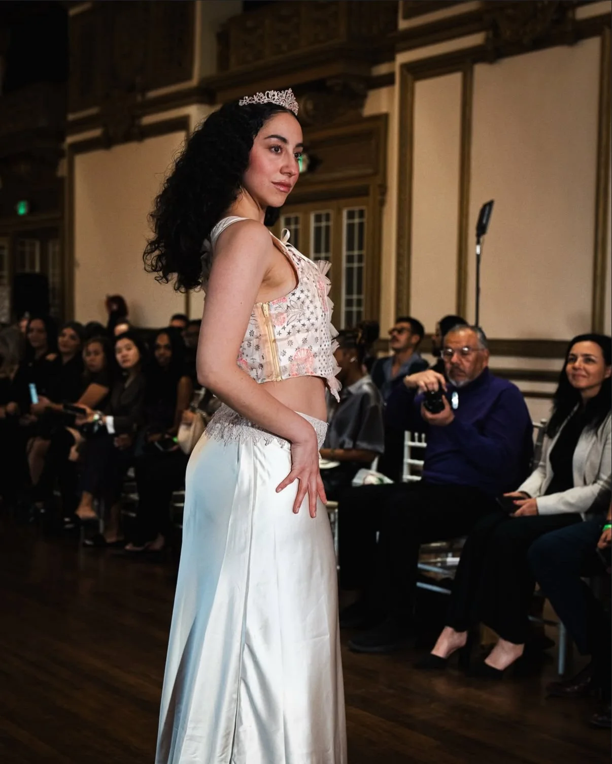 A woman wearing a tiara, a floral crop top, and a long satin skirt stands in front of an audience at an indoor event.