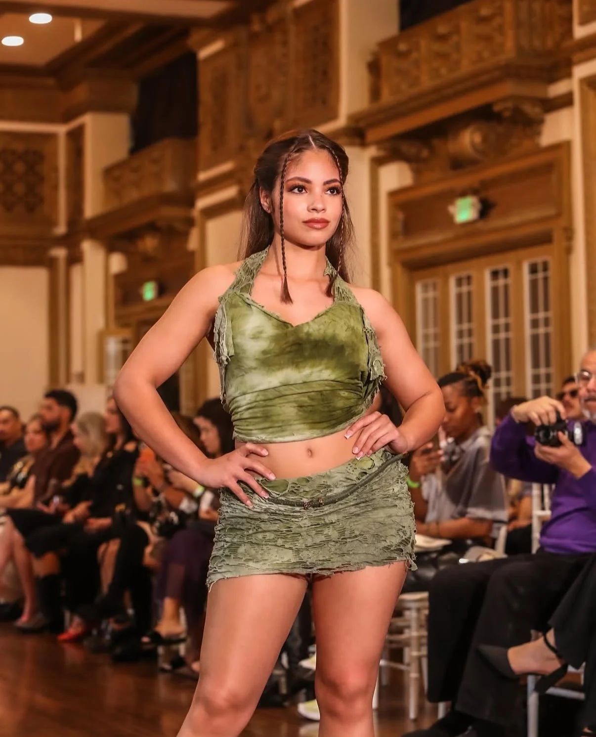 Young woman walking on a runway in a green tie-dye tank top and matching distressed mini skirt during a fashion show.