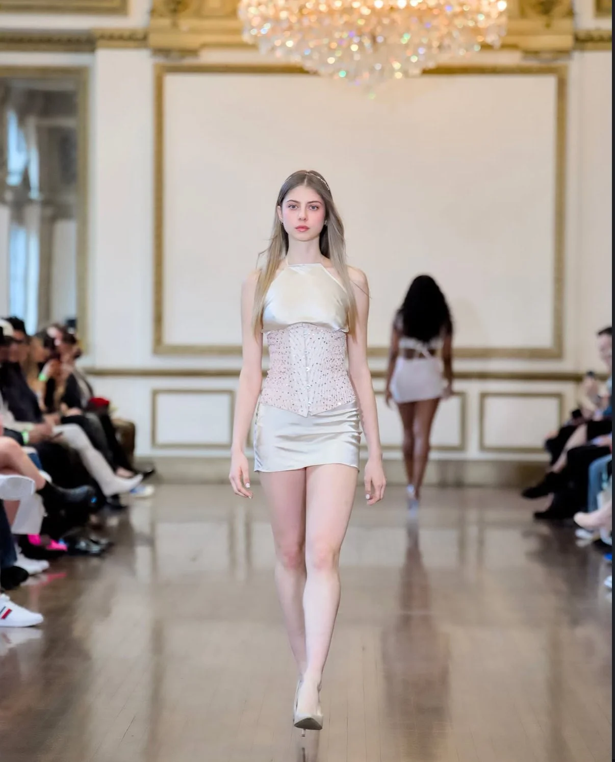 A female model walking down a runway during a fashion show, wearing a cream-colored outfit with a sleeveless top and shorts, in a luxurious room with gold accents and a chandelier.