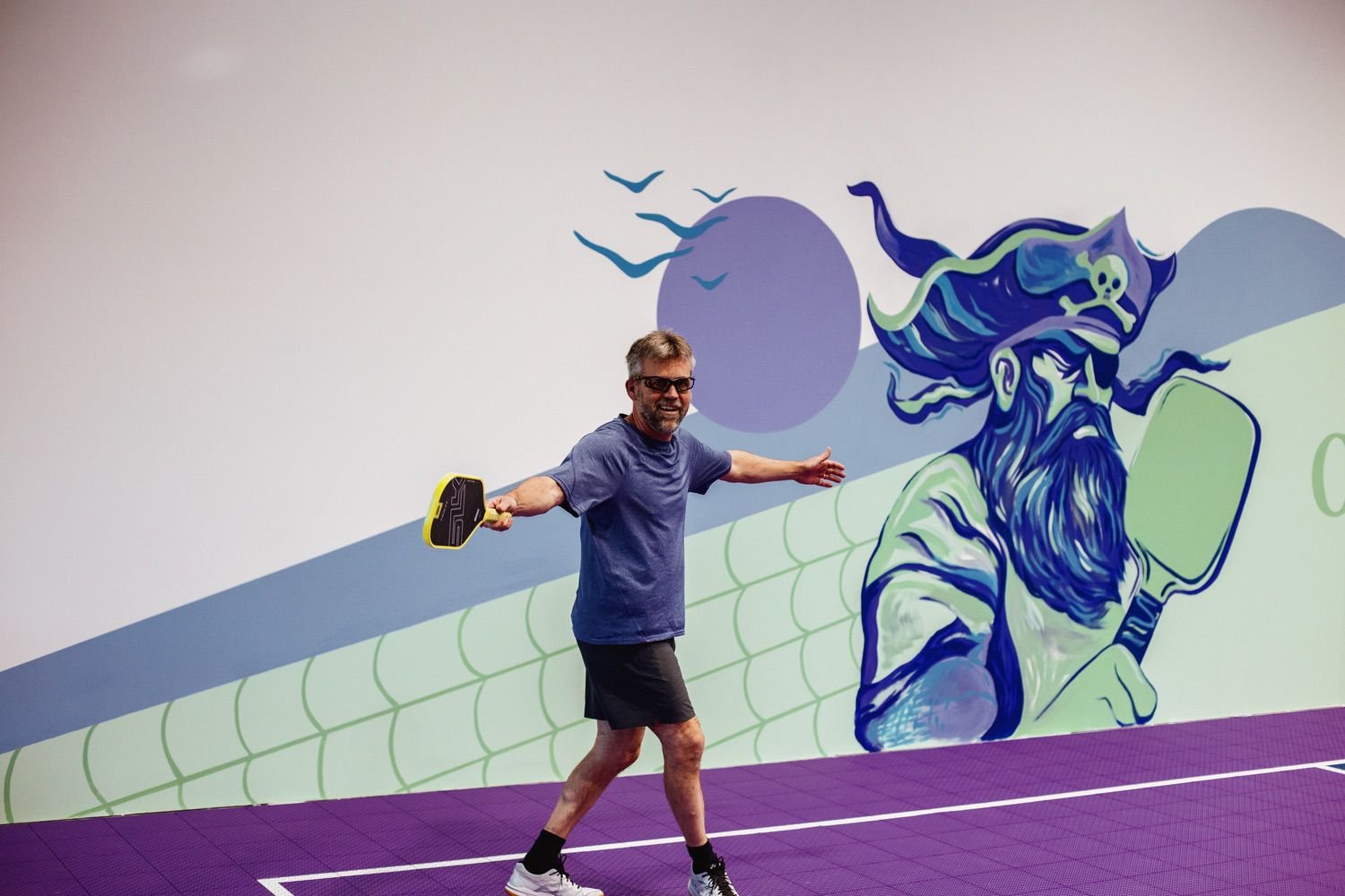 Man in gray shirt and black shorts playing pickleball on purple court with a colorful mural of a wizard in the background.