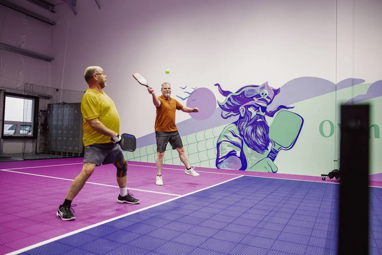 Two men playing pickleball on an indoor court with purple and blue flooring, one with a yellow shirt and the other with an orange shirt, in front of a wall with colorful mural art of a bearded pirate holding a paddle.