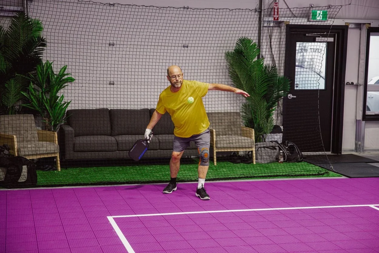 Man playing pickleball indoors on a purple court, wearing a yellow shirt, shorts, and sneakers, holding a paddle, with a net and plants in the background.
