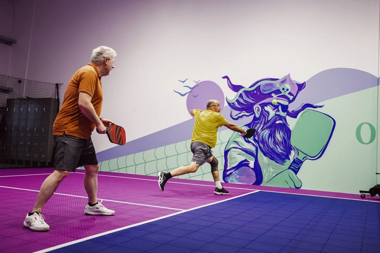 Two elderly men playing pickleball indoors on a pink and blue court with a colorful mural of a bearded wizard on the wall.