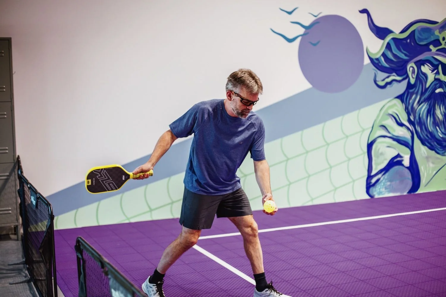 A man playing pickleball on an indoor court with purple flooring, holding a paddle in one hand and a ball in the other, in front of a mural with a dragon and a large moon.