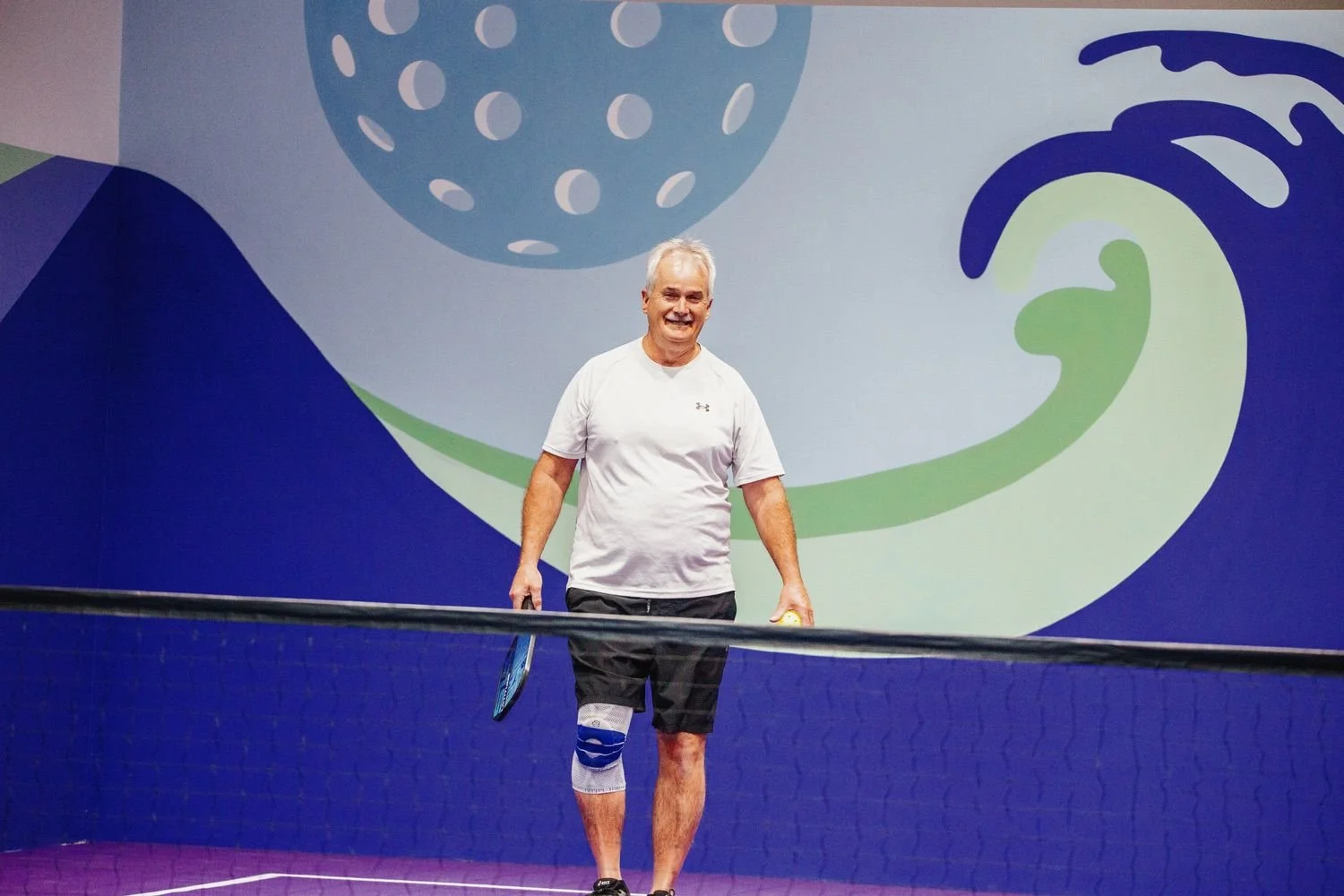 older man playing pickleball on an indoor court with a purple floor, holding a paddle, wearing a knee brace, smiling, with a colorful abstract mural in the background.
