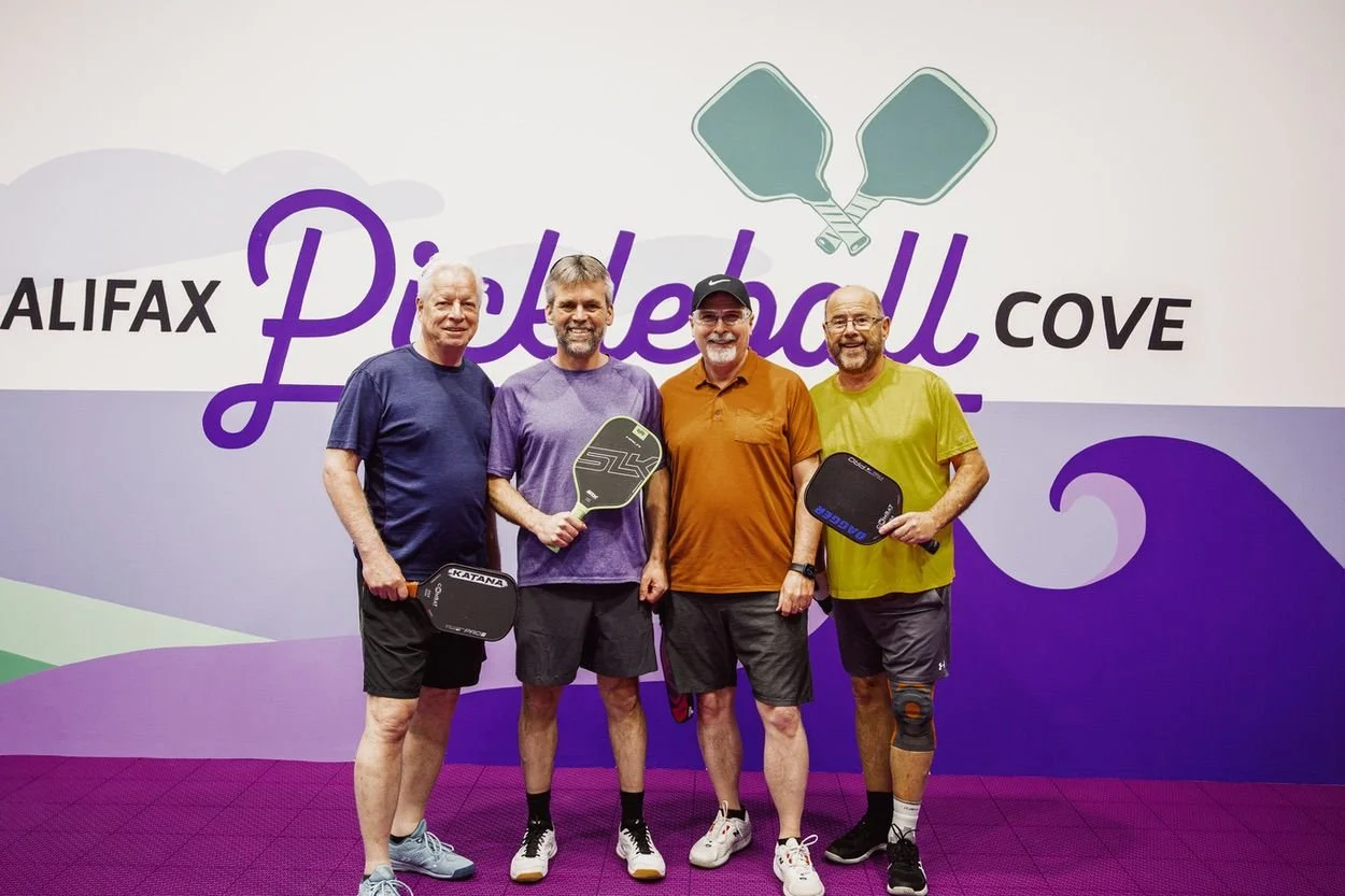 Four men standing in front of a backdrop that reads HALIFAX Pickleball COVE' with pickleball paddles, smiling.