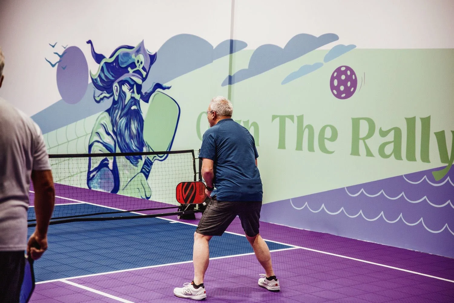People playing pickleball in an indoor court with bright purple and blue flooring. A large mural on the wall depicts a pirate with a beard, a hat with a skull, and a rainbow in a cartoon style. Part of a banner with the words "On The Rally" is visible.