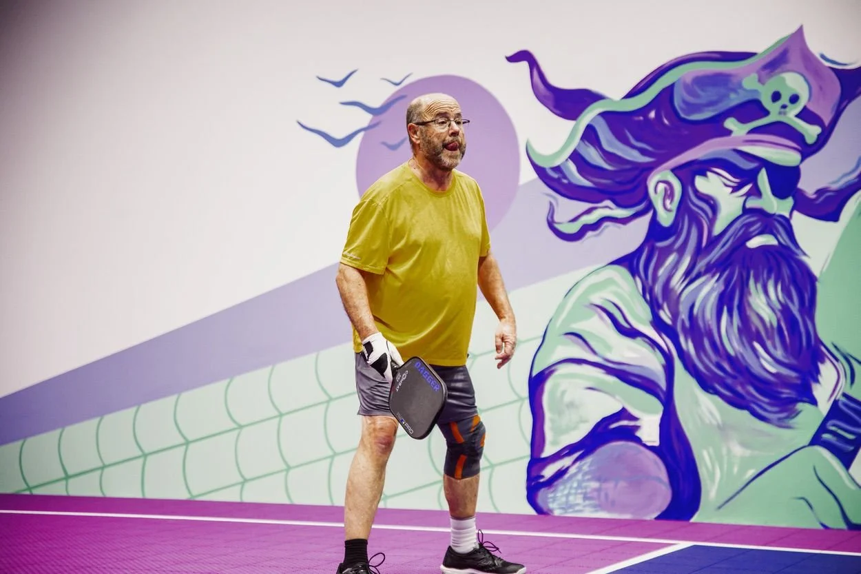 An older man with glasses, in a yellow t-shirt and shorts, holding a pickleball paddle, standing on a purple pickleball court with a purple and green dragon mural in the background.
