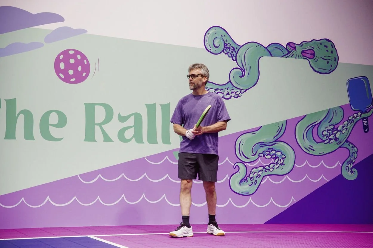 A man in athletic clothing holding a pickleball paddle on a pickleball court with a colorful, cartoon-style mural background.