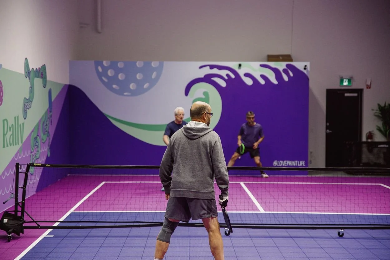 People playing pickleball on an indoor court with colorful wall art.