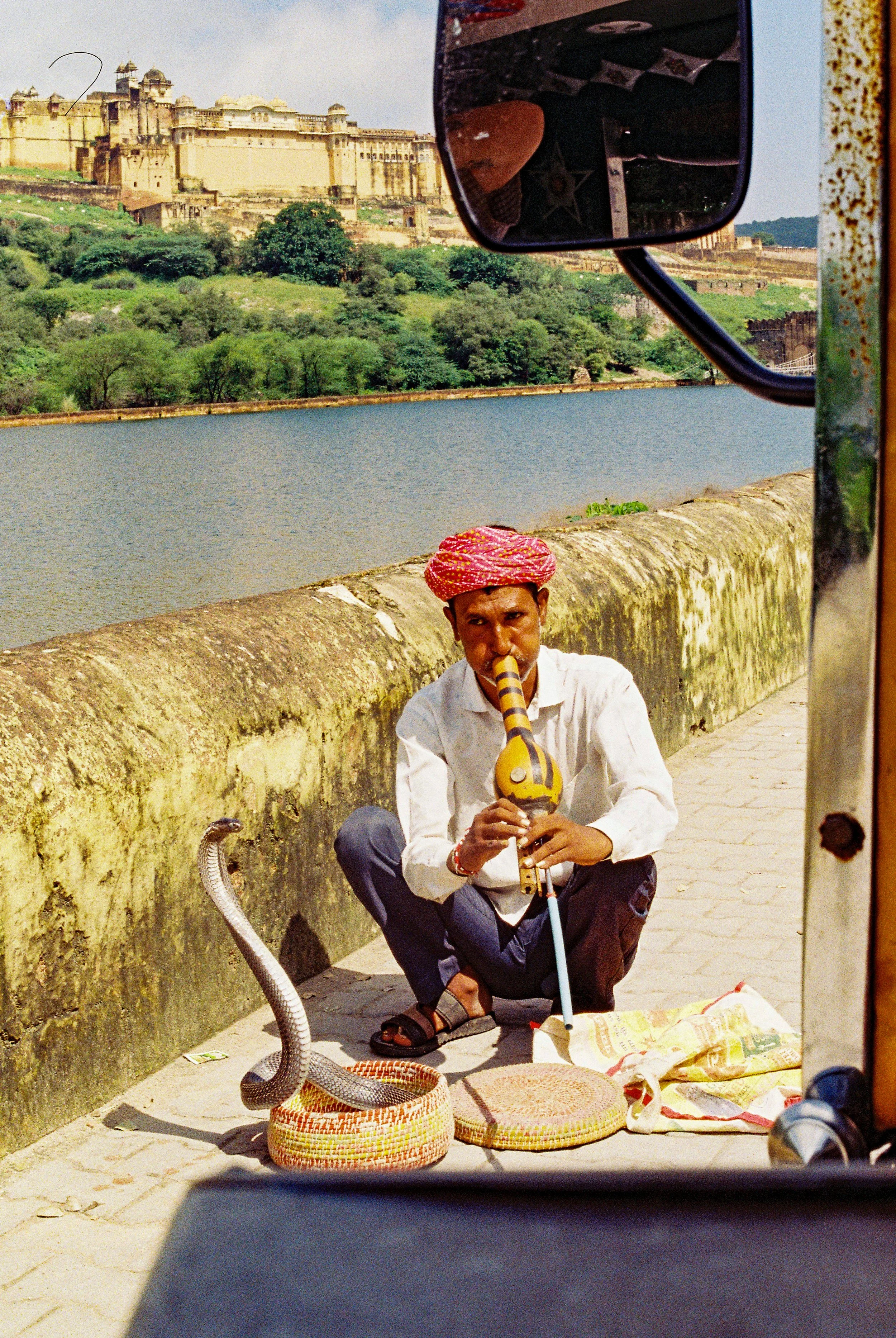 A man wearing a pink turban and white shirt sitting on the ground, playing a traditional Indian flute, with a lake and a historic fort on a hill in the background. 35mm film photography. Kodak Ultramax 400. Fujifilm Frontier SP-3000 high-resolution s