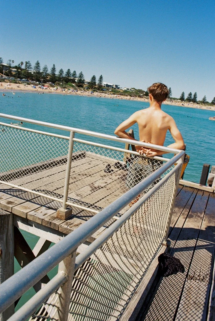 A boy with short hair and no shirt standing on a wooden pier overlooking the ocean in Port Elliot, South Australia, with a sandy beach and trees in the background on a sunny day. 35mm film photography. Kodak Gold 200. Fujifilm Frontier SP-3000 high-r