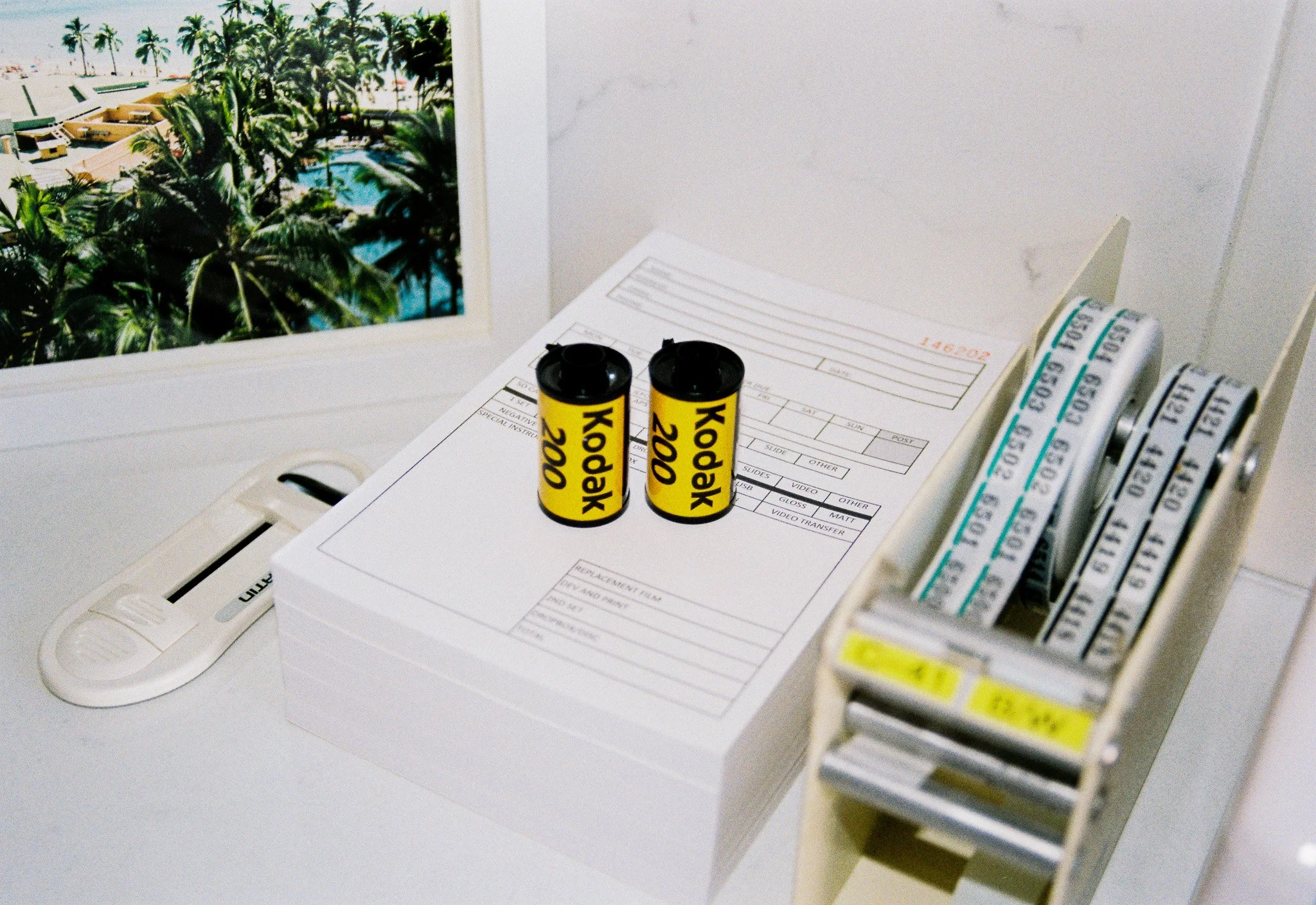 Office desk with two yellow Kodak 200 film canisters, a stack of film rolls, and a paper with forms. There is a desktop tray in the left foreground and a monitor displaying a tropical beach scene with palm trees and a swimming pool in the background.