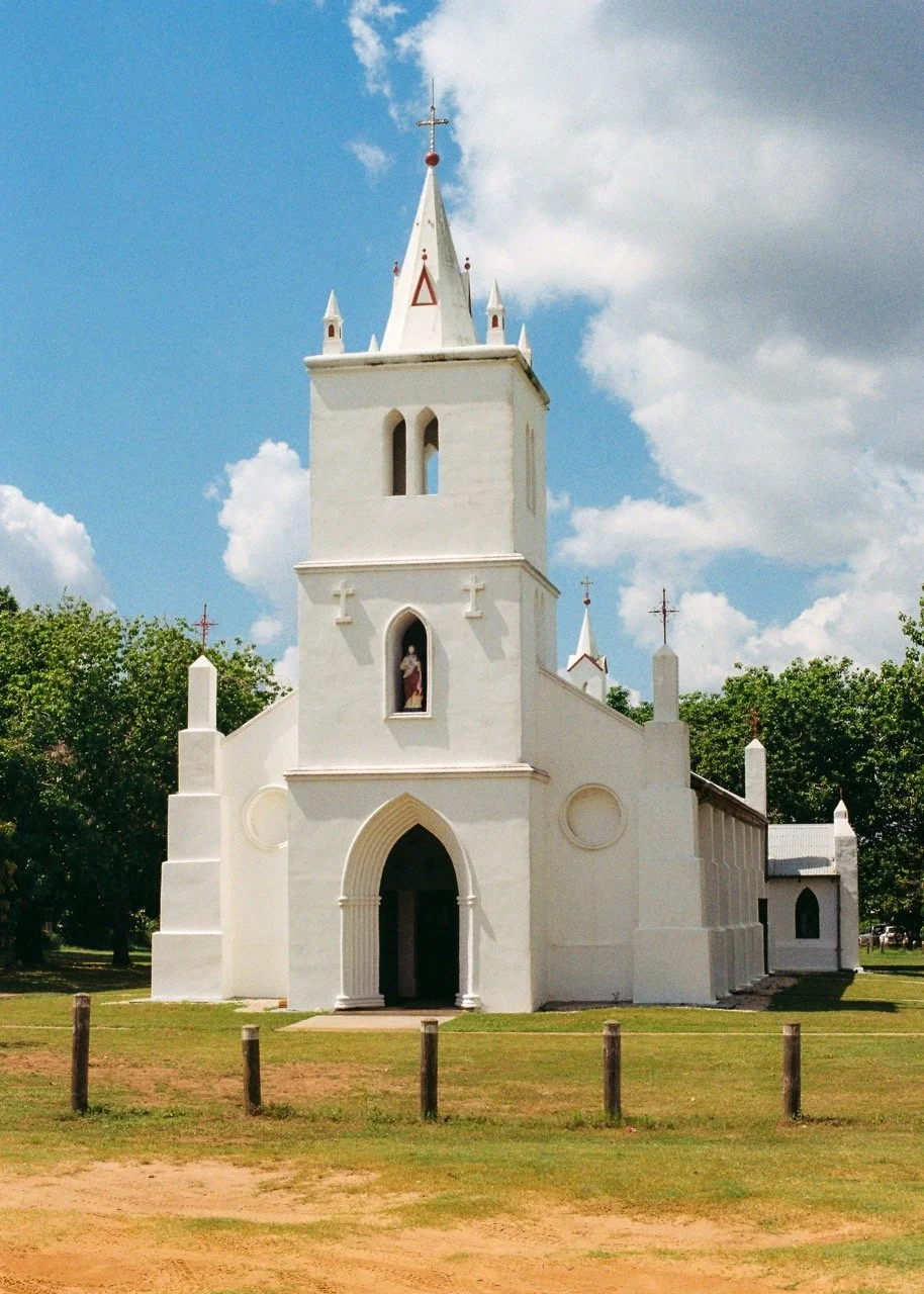 A white church with a tall steeple and cross at the top, surrounded by trees and a grassy area, under a partly cloudy sky. 35mm film photography. Kodak Ultramax 400. Fujifilm Frontier SP-3000 high-resolution scan. Professional mail-in film processing
