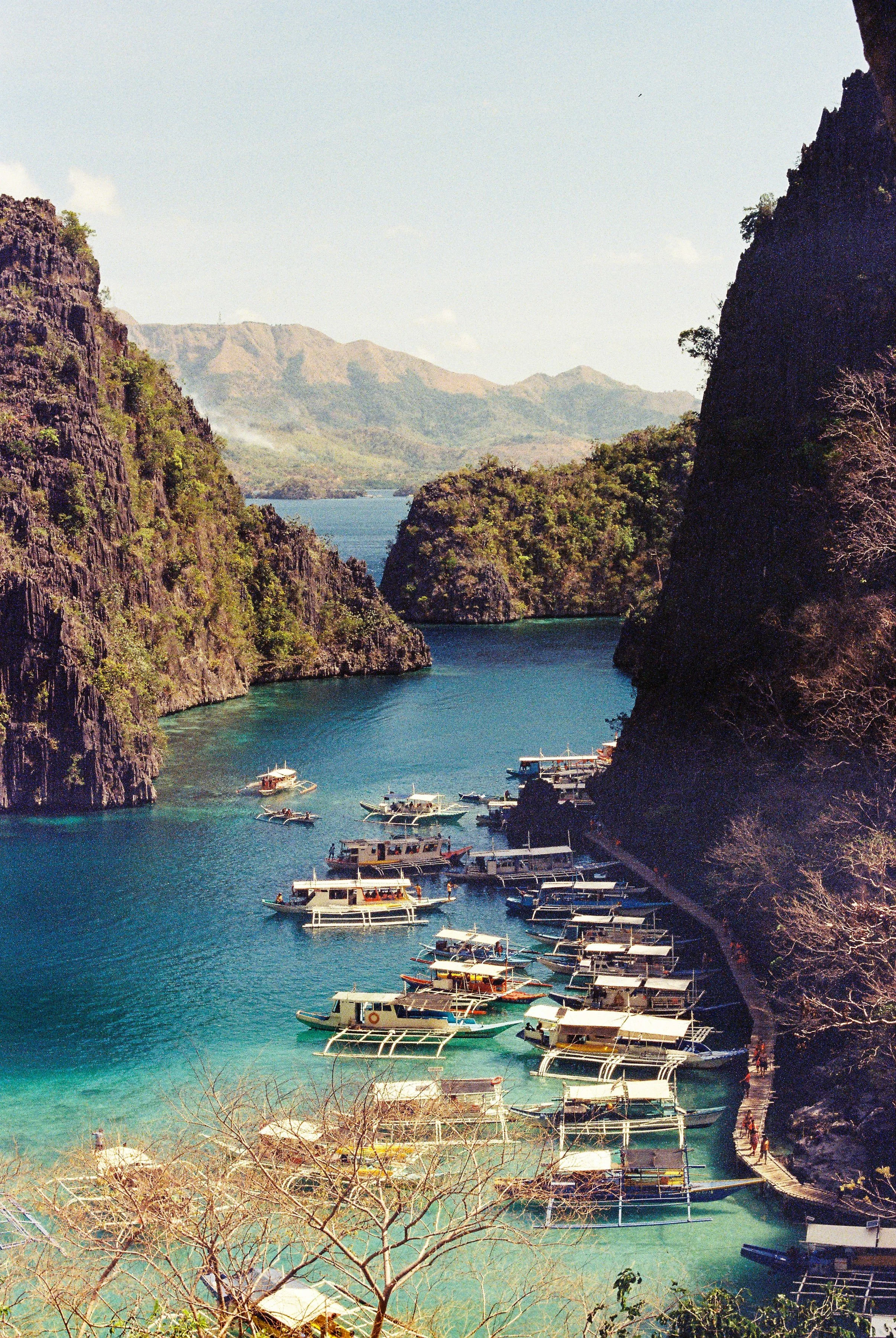 A scenic view of a cove with turquoise waters, surrounded by steep rocky cliffs with greenery, and several boats docked or sailing near a small pier, with mountains and sparse clouds in the background. 35mm film photography. Kodak Ultramax 400. Fujif