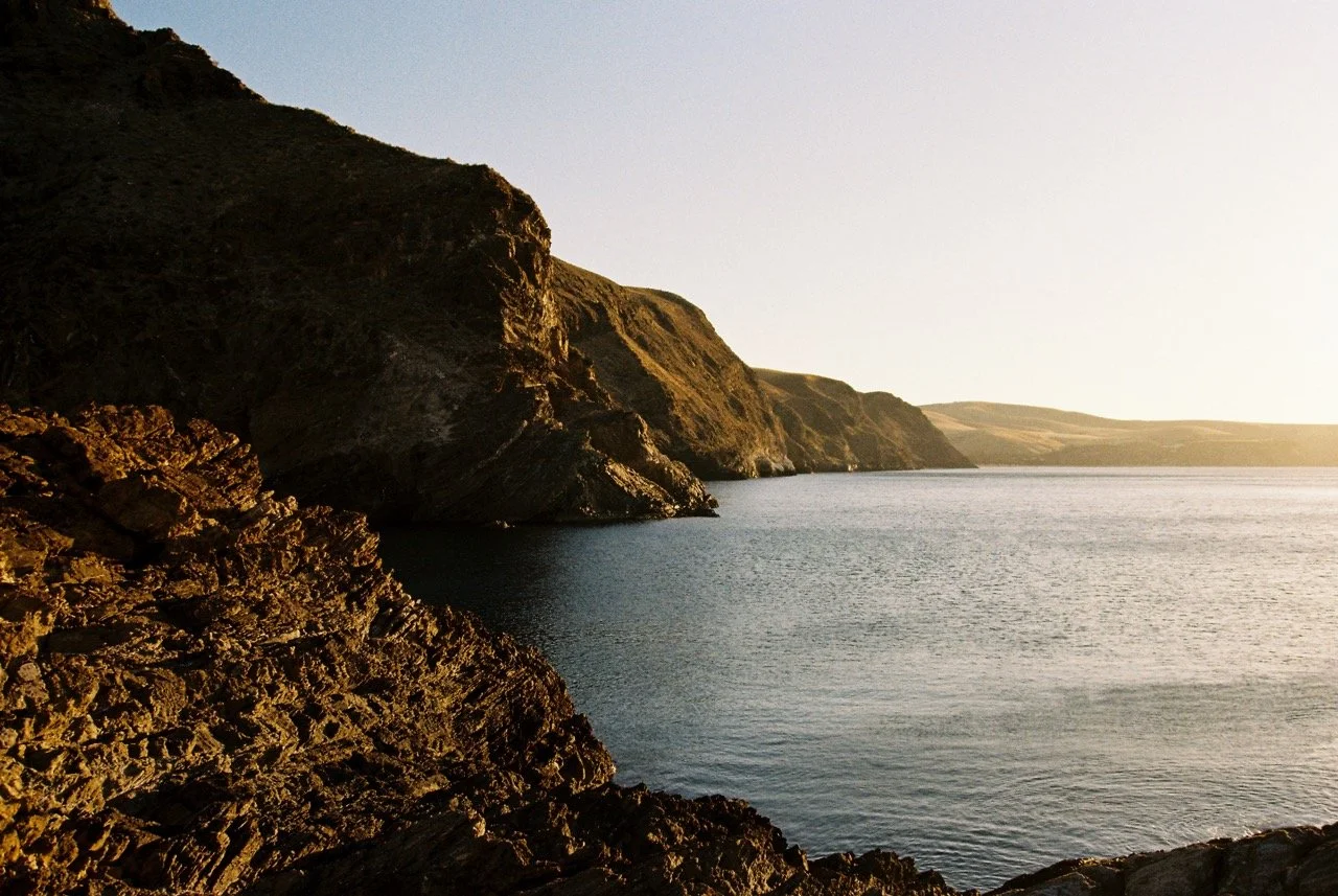 A rocky coastline with cliffs on the left and calm water extending to the horizon, with the sun setting or rising in the distance. Second Valley South Australia. Nikon F80 film camera. 35mm film photography. Kodak Gold 200. Fujifilm Frontier SP-3000 