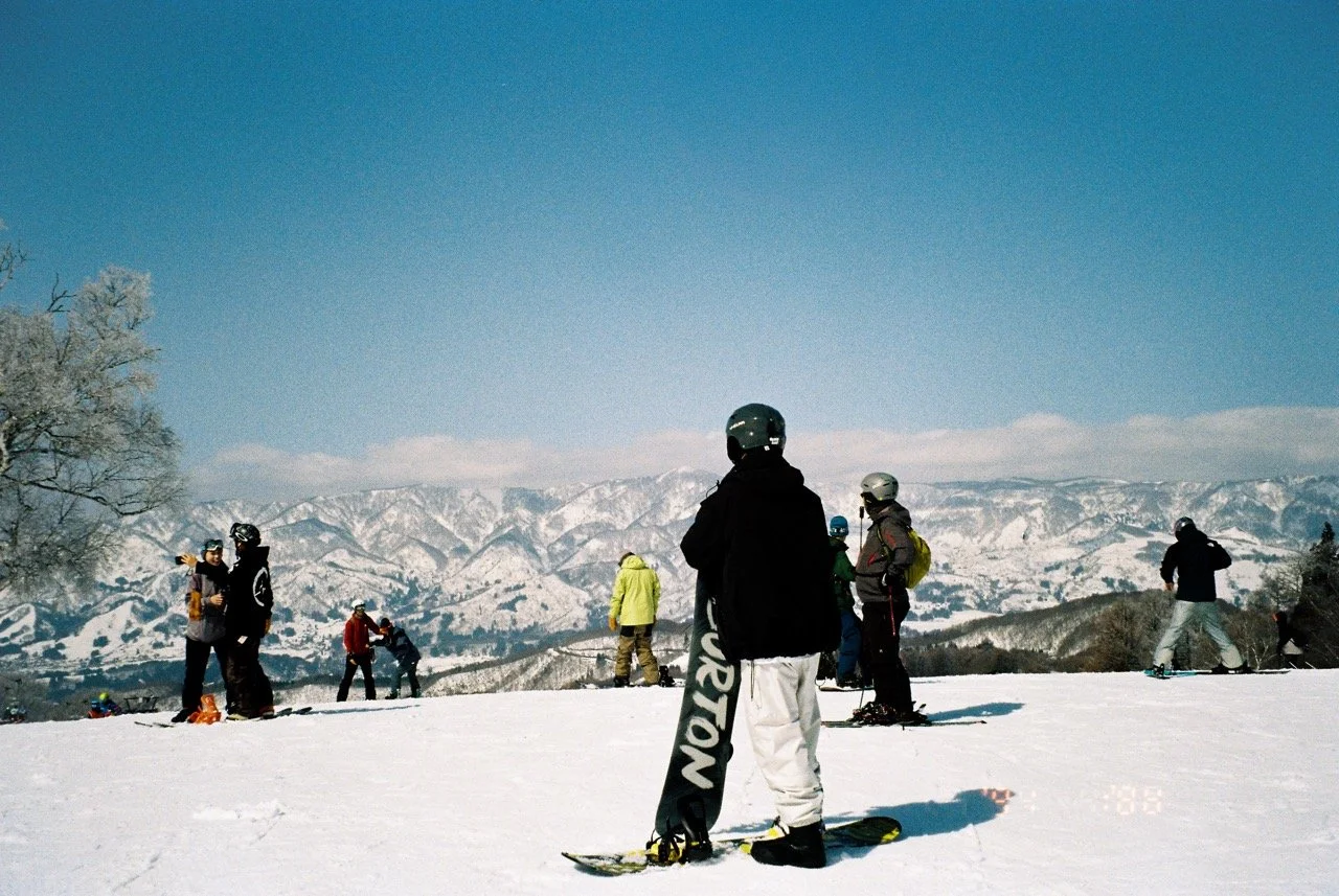 A group of people snowboarding and skiing on a snowy mountain in Japan with a clear blue sky and snow-covered mountains in the background. 35mm film photography. Kodak Ultramax 400. Point and shoot camera. Olympus MJU. Flash film photography. Fujifil