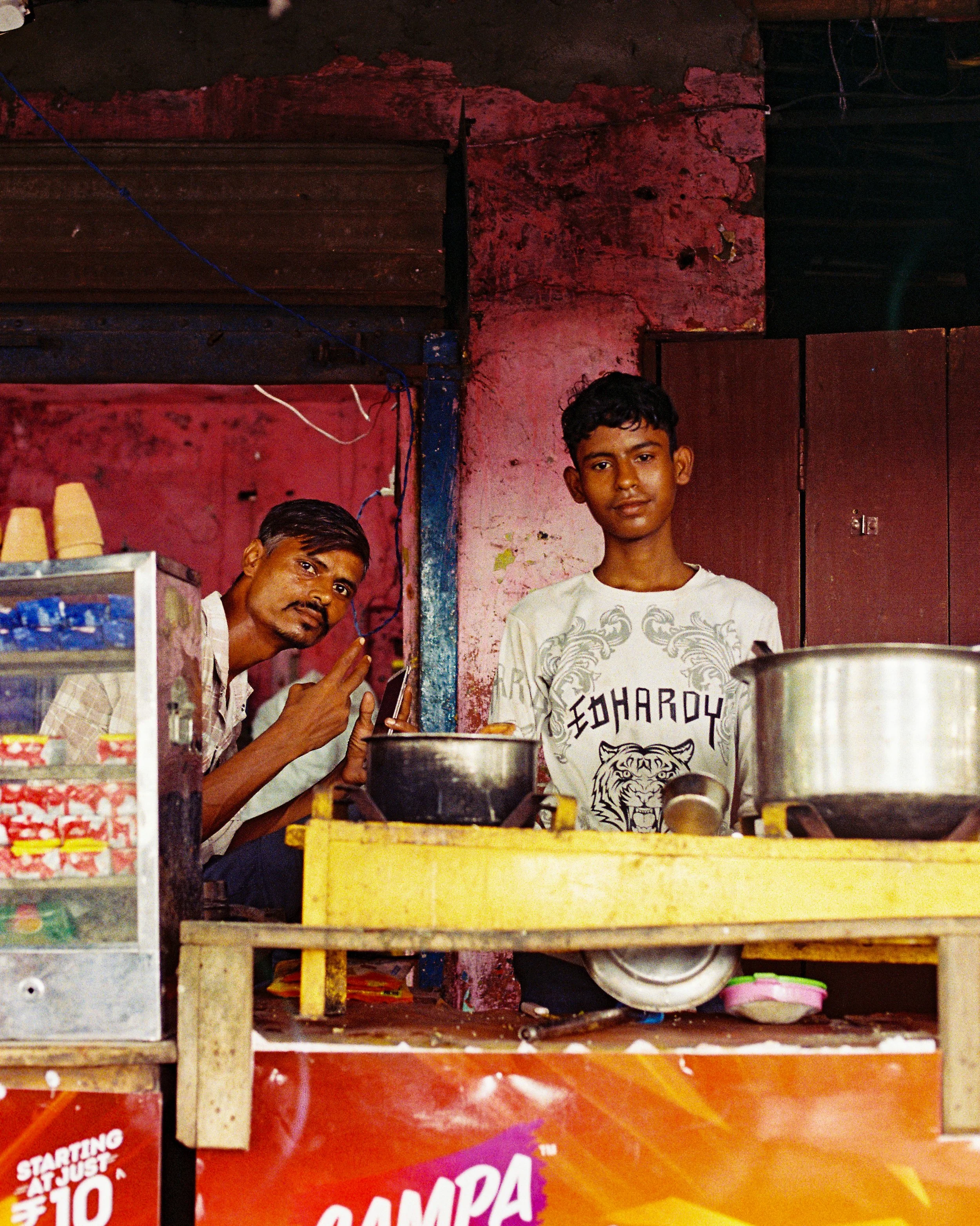 Two young men standing behind a colorful store counter in a corner of a shop. The store has pink and red painted walls, and shelves with various items for sale, including snacks and beverage bottles. One man is wearing a white t-shirt with a tiger gr