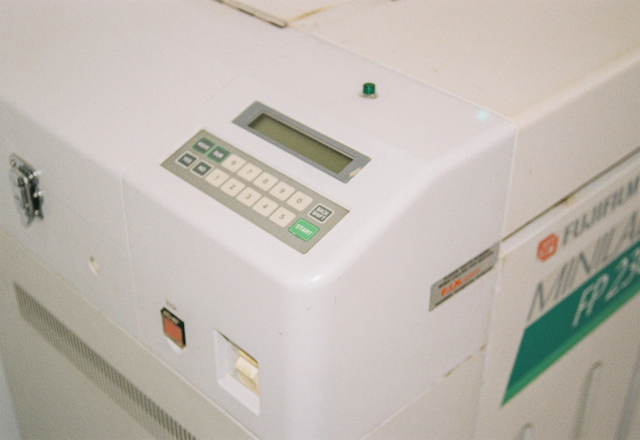 A white electronic safe with a keypad and display on top, placed on a flat surface, with some labels and signs on its side.