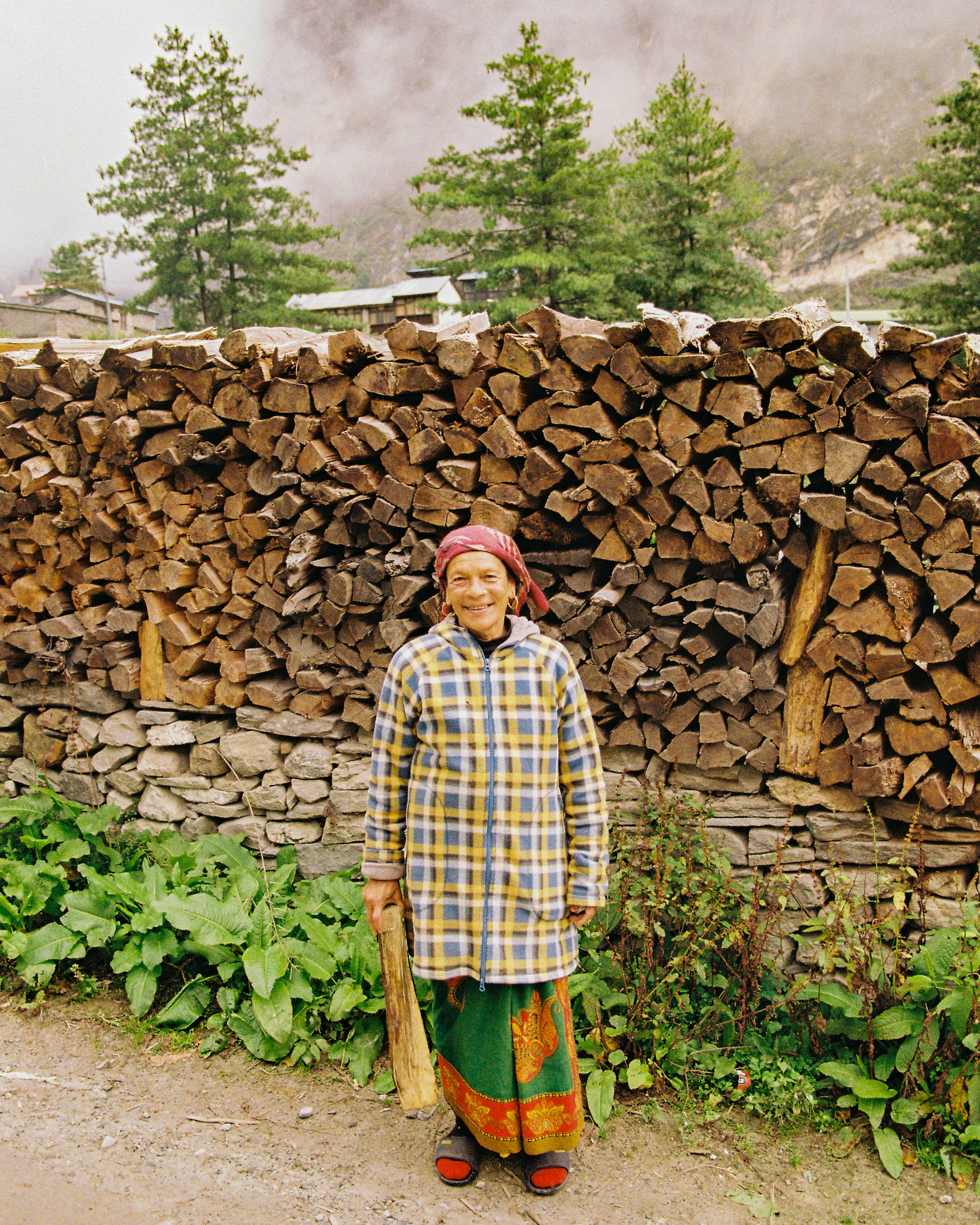 A person in traditional attire standing outdoors in Peru in front of a large stack of chopped firewood and green plants. The person is smiling and holding a stick. 35mm film photography. Kodak Ultramax 400. Fujifilm Frontier SP-3000 high resolution s