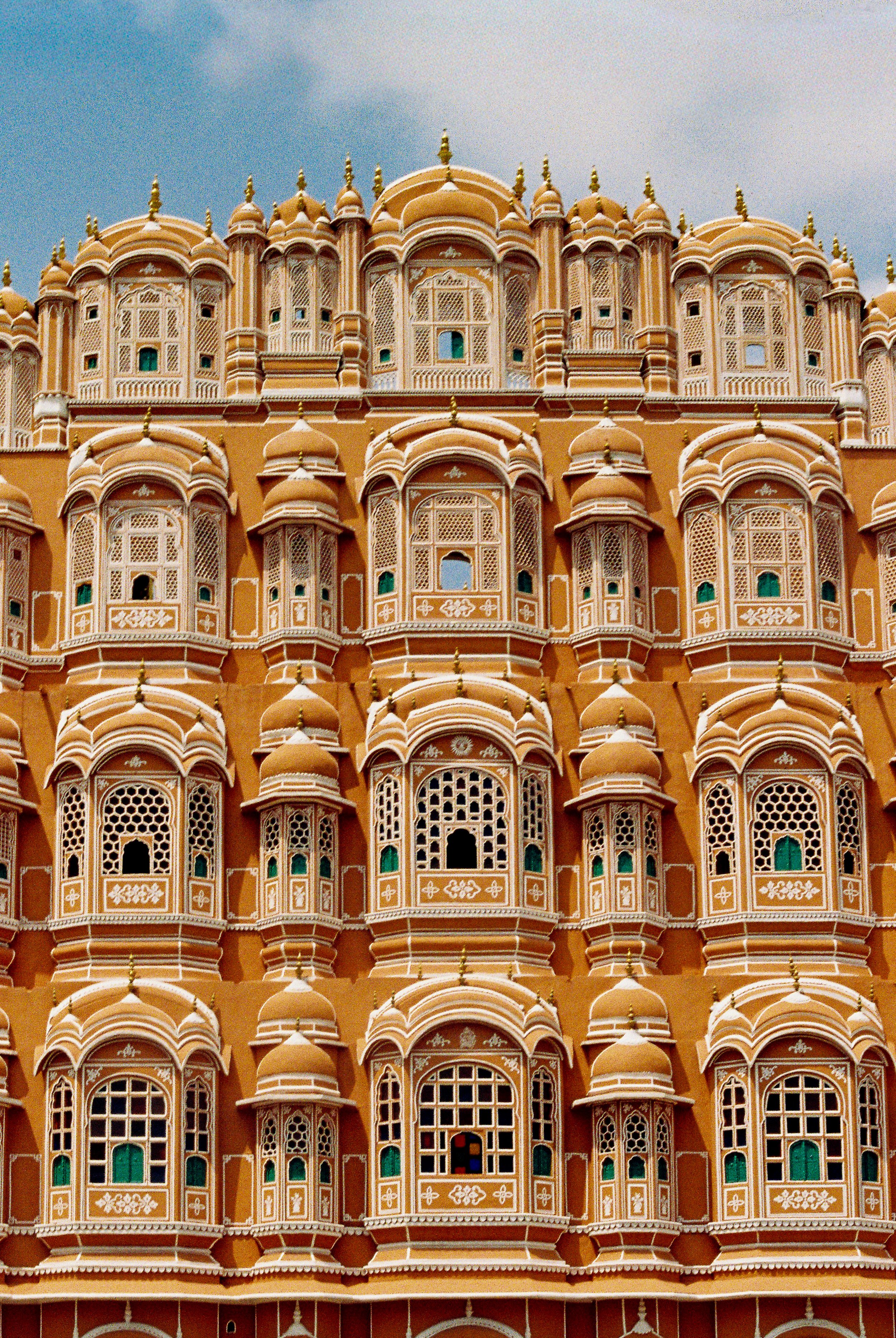 Close-up of the intricate architecture and detailed windows of the Hawa Mahal building in Jaipur, India. The facade features multiple small windows with ornate lattice work and domed structures in a pink color. 35mm film photography. Kodak Ultramax 4