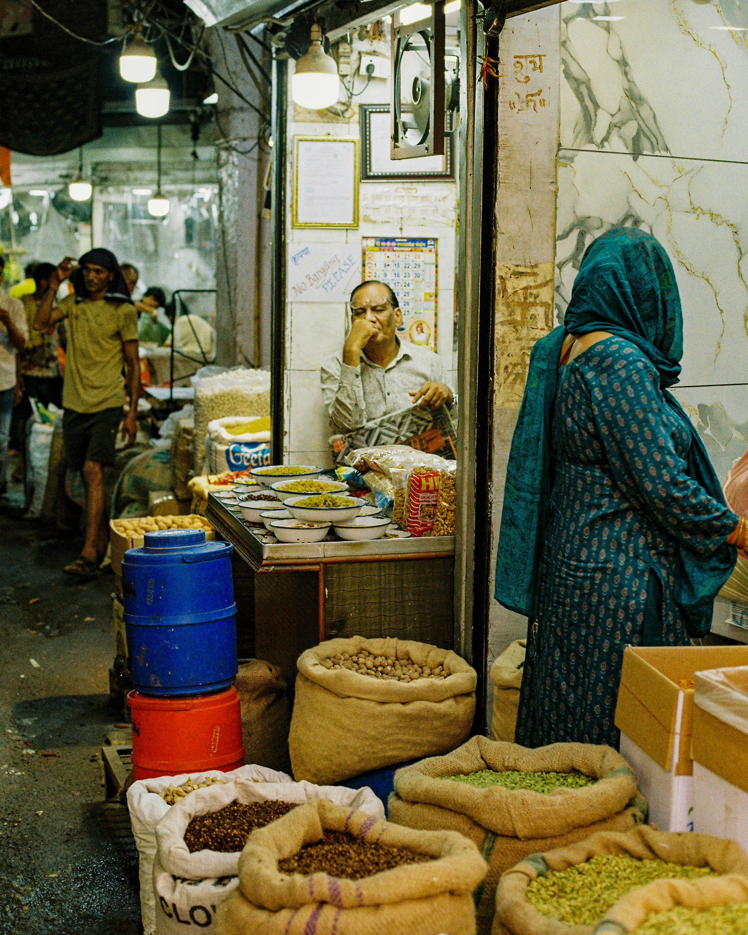 A busy marketplace with various sacks and containers filled with different grains, spices, and dried goods. People are walking and shopping, with some standing near the display of food items. Bright lights are hanging overhead, and the scene is vibra