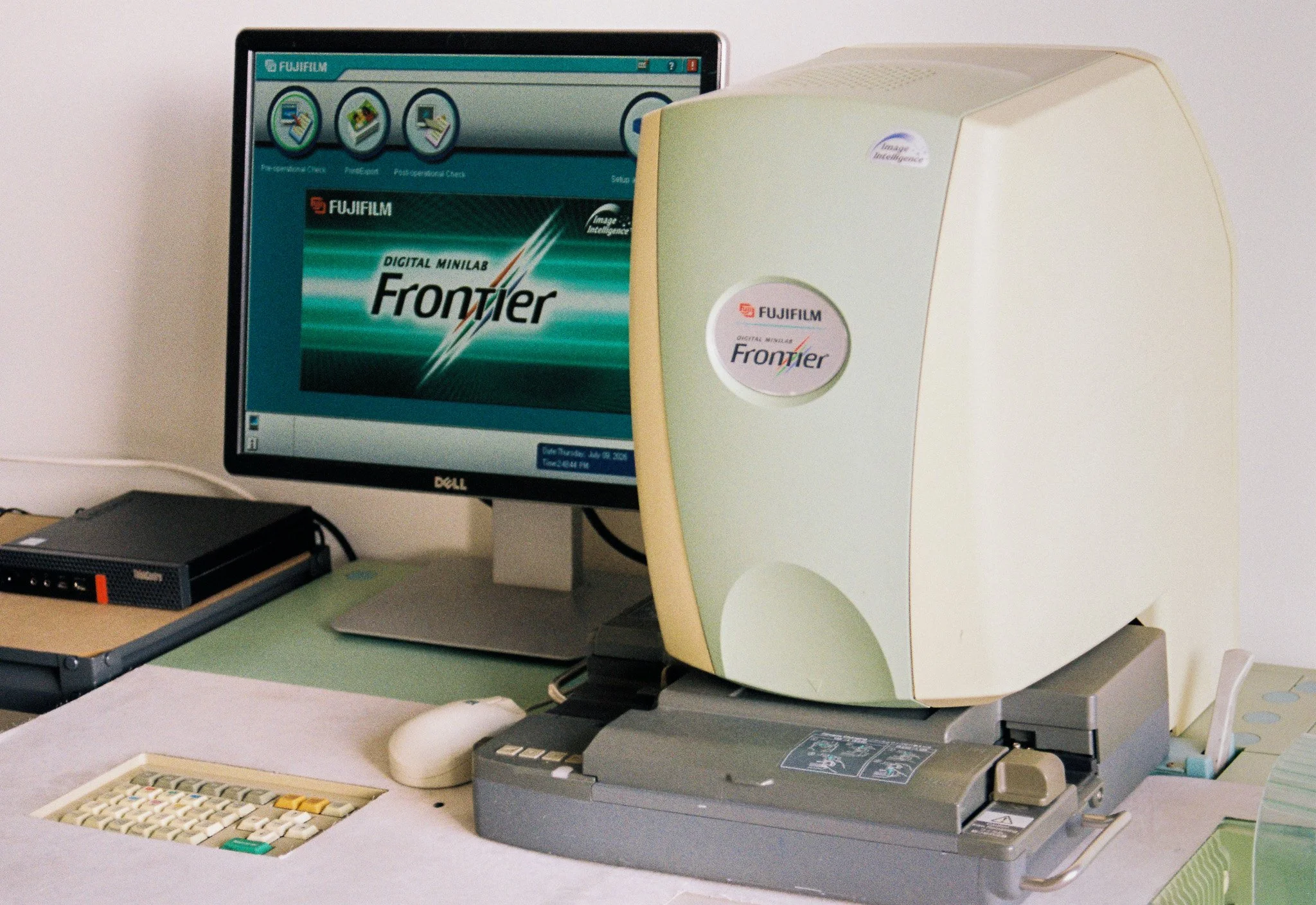Older computer monitor with a desktop screen displaying a Fujifilm software interface, and a dot matrix printer on a desk, with an open box and color swatch samples nearby.