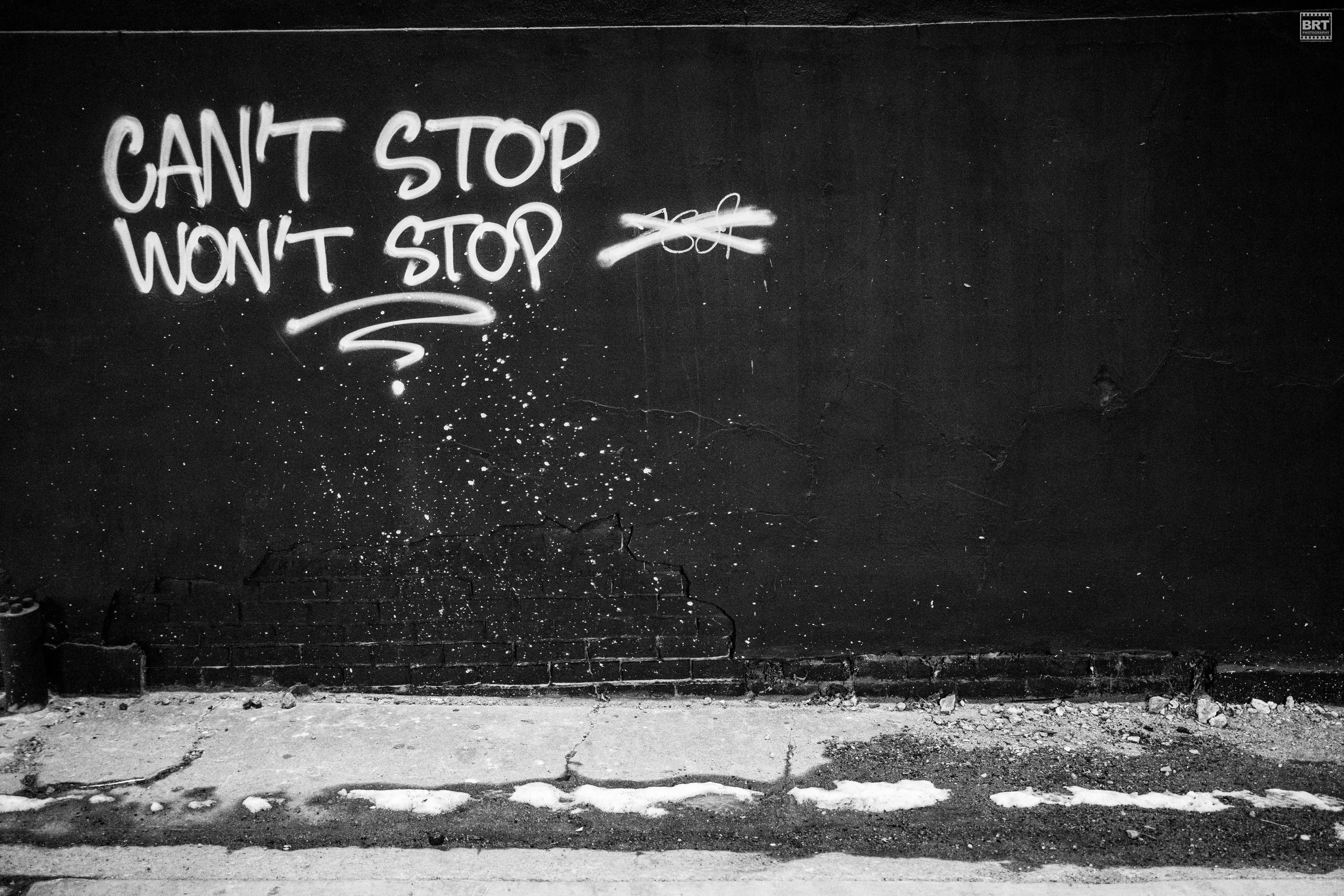 Black wall with graffiti stating 'Can't stop, won't stop' in white spray paint, with some paint splatters and cracks on the wall, and a concrete ground with patches of snow or paint.
