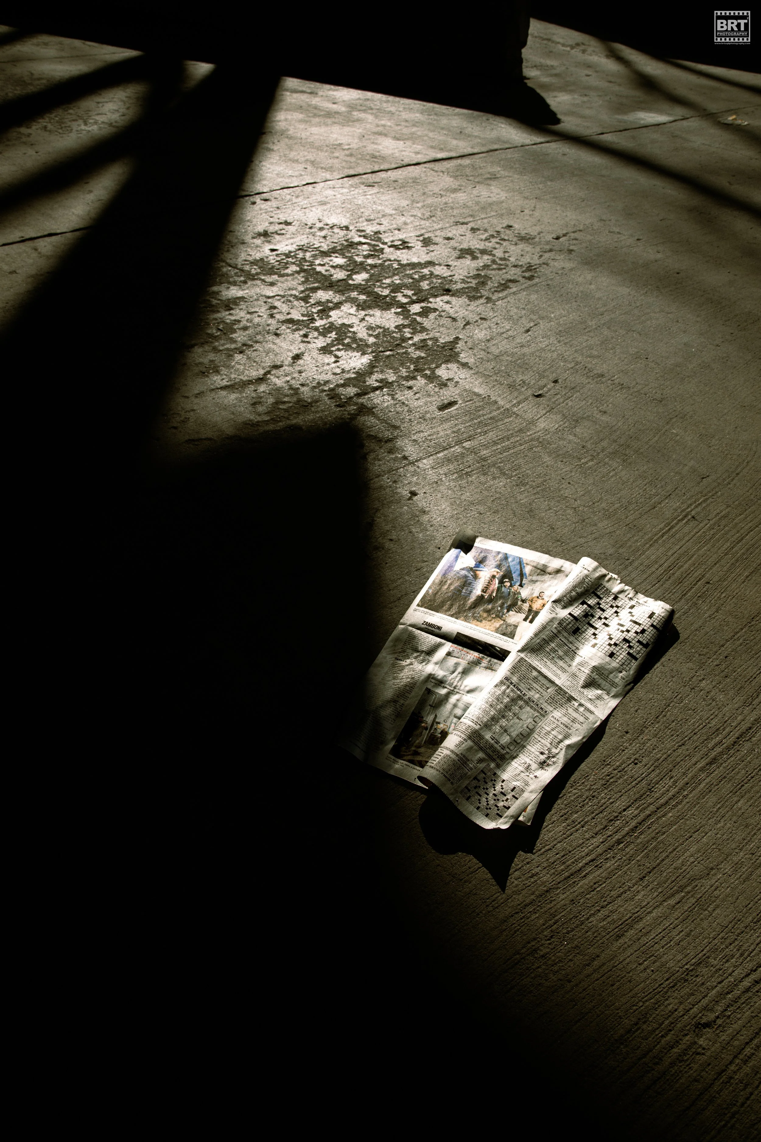 A newspaper lying on a concrete floor partially in shadow, with sunlight creating stark contrast and shadows.