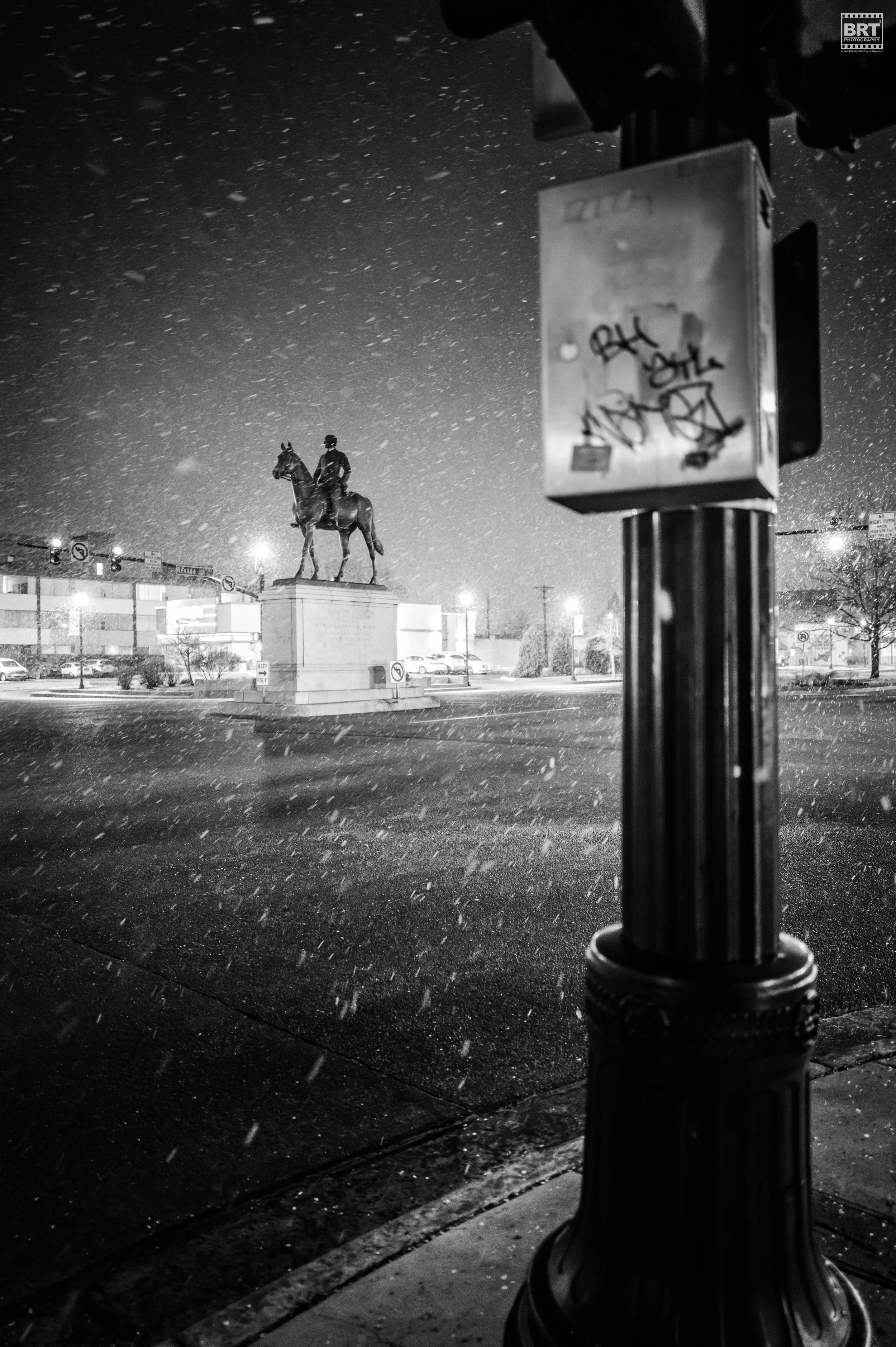 A snowy night scene featuring an equestrian statue of a man on a horse, illuminated and surrounded by falling snow, with a street corner and traffic lights in the background. In the foreground, there is a street pedestrian button and a pole with graf