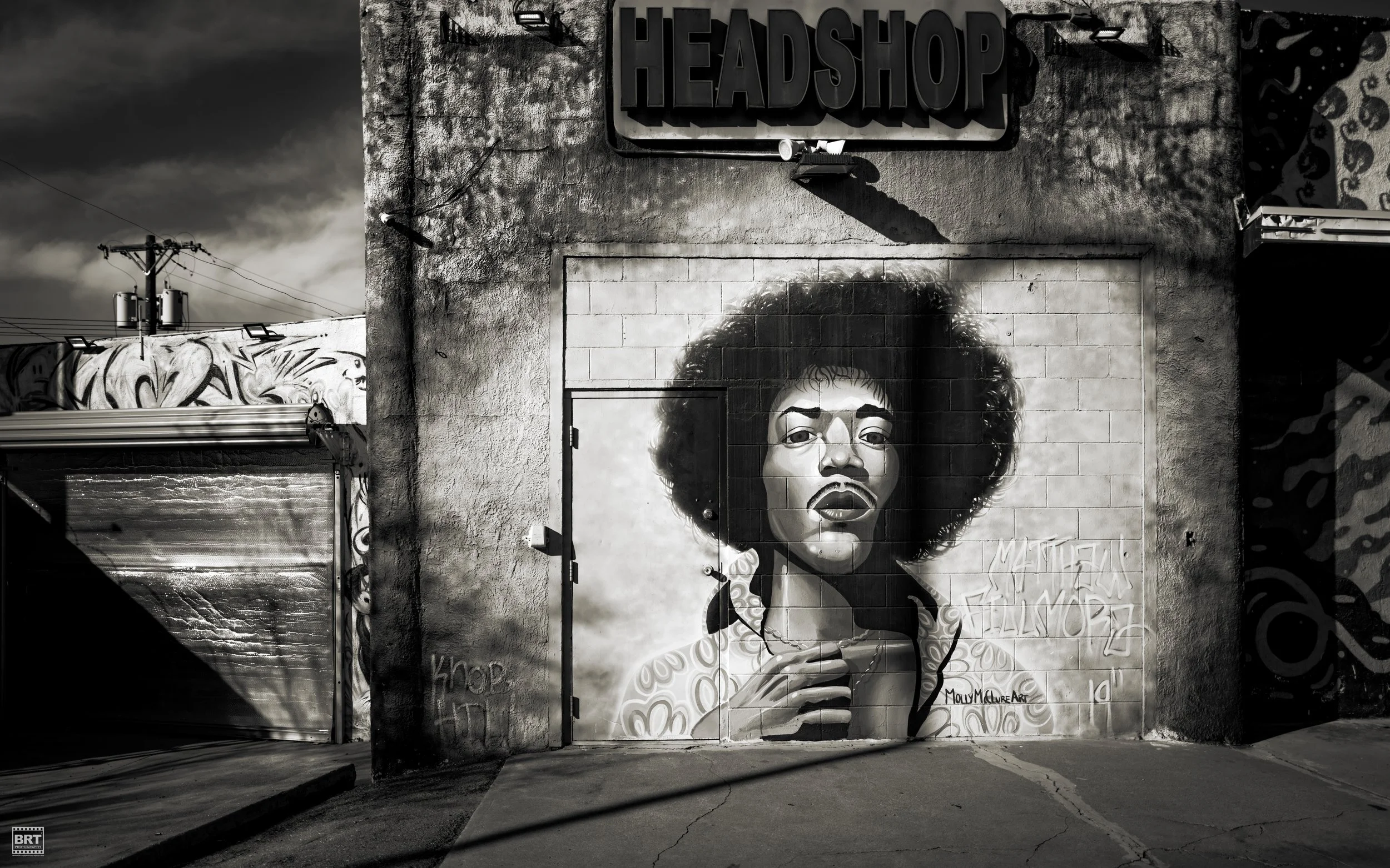 Black and white mural of a woman with curly hair on a building wall, with a sign above that reads 'HEADSHOP'.