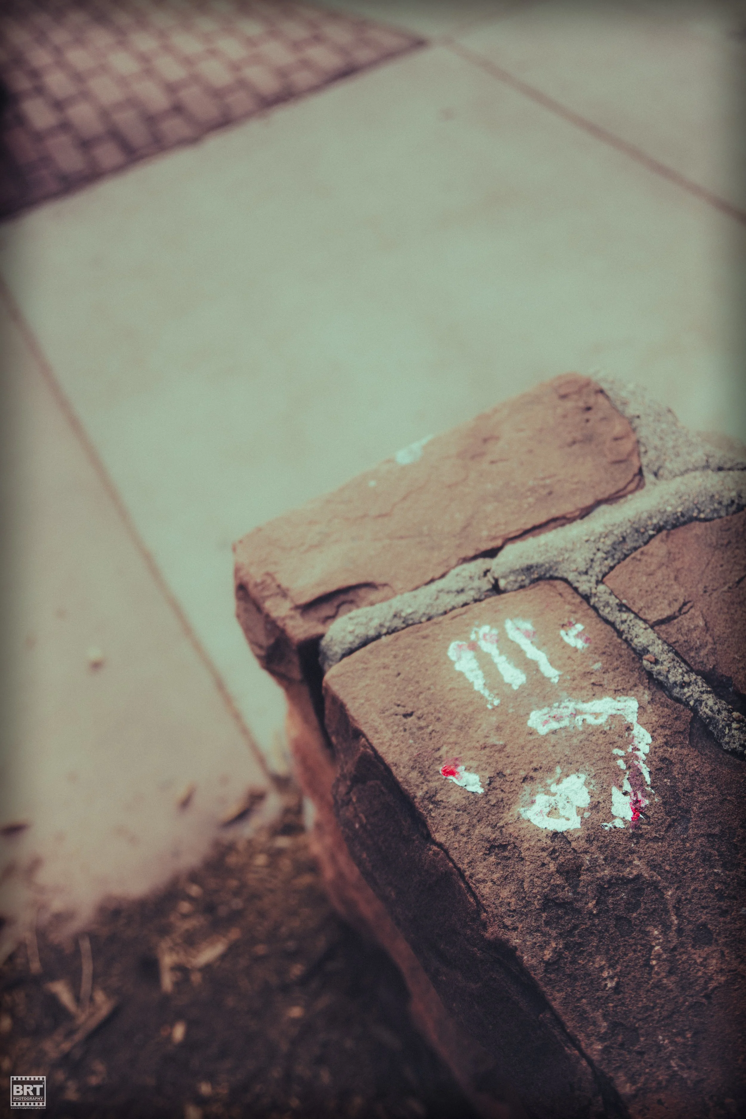 Close-up of a weathered brick with white chalk graffiti of a smiley face and three vertical lines, on a brick wall. The background shows a sidewalk with beige tiles and a small section of reddish-brown paving stones.