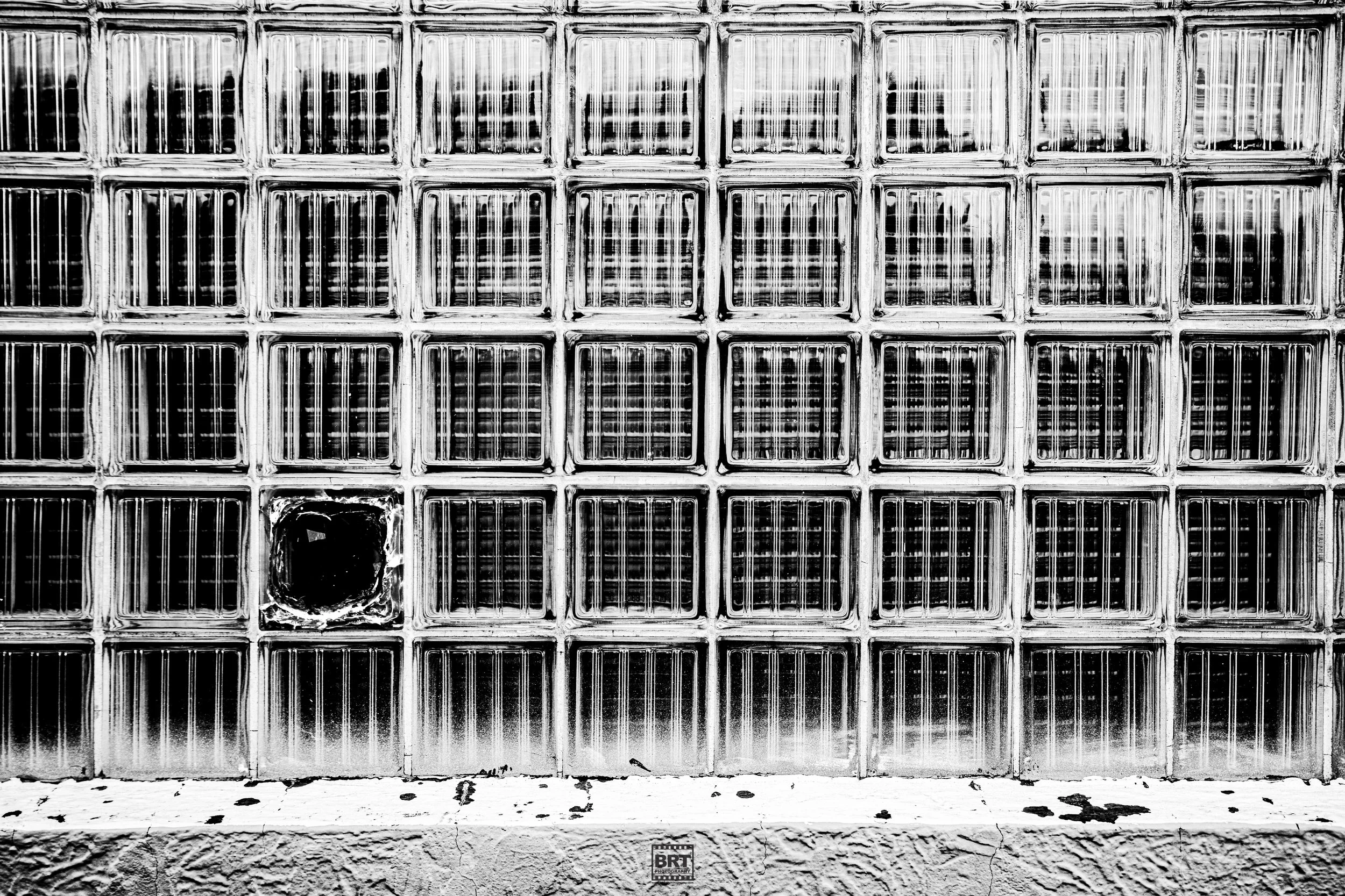 A wall made of glass bricks with one brick that is broken and missing, in black and white.