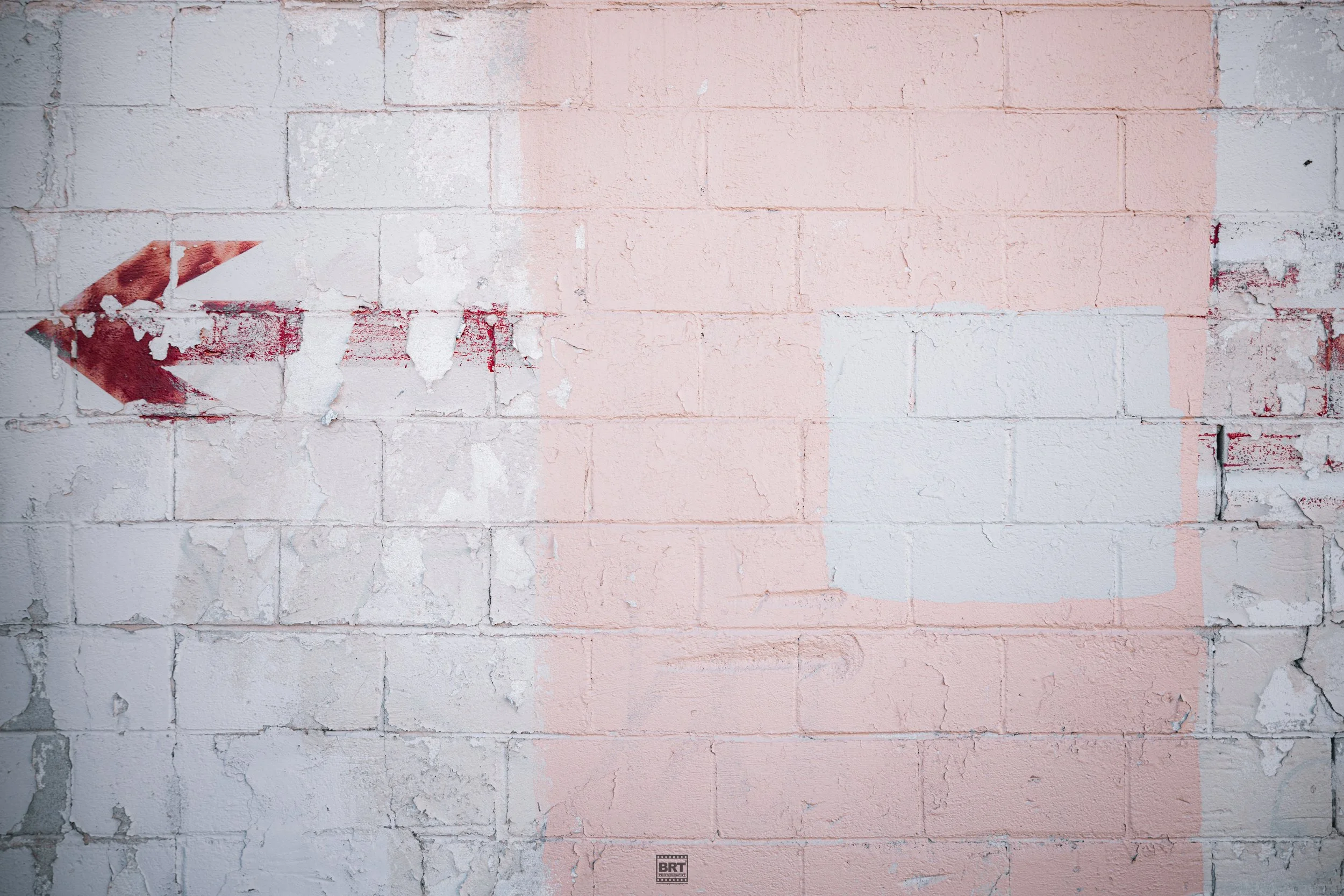 White brick wall with a red arrow painted on it, pointing to the left, with some peeling paint and patches of pink paint.