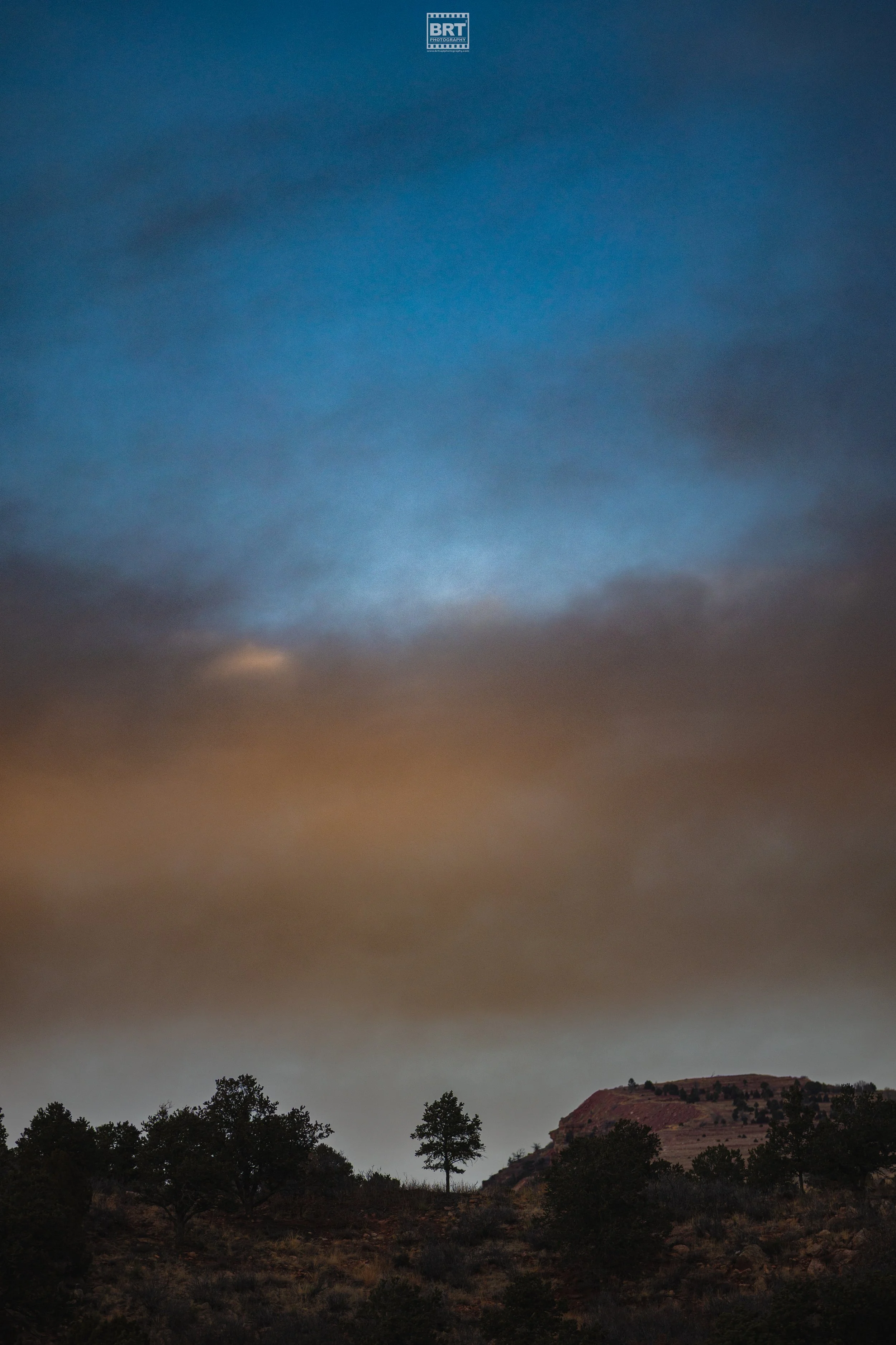 A landscape photo of trees on a hill with a rocky formation, under a cloudy sky that transitions from dark gray to blue.