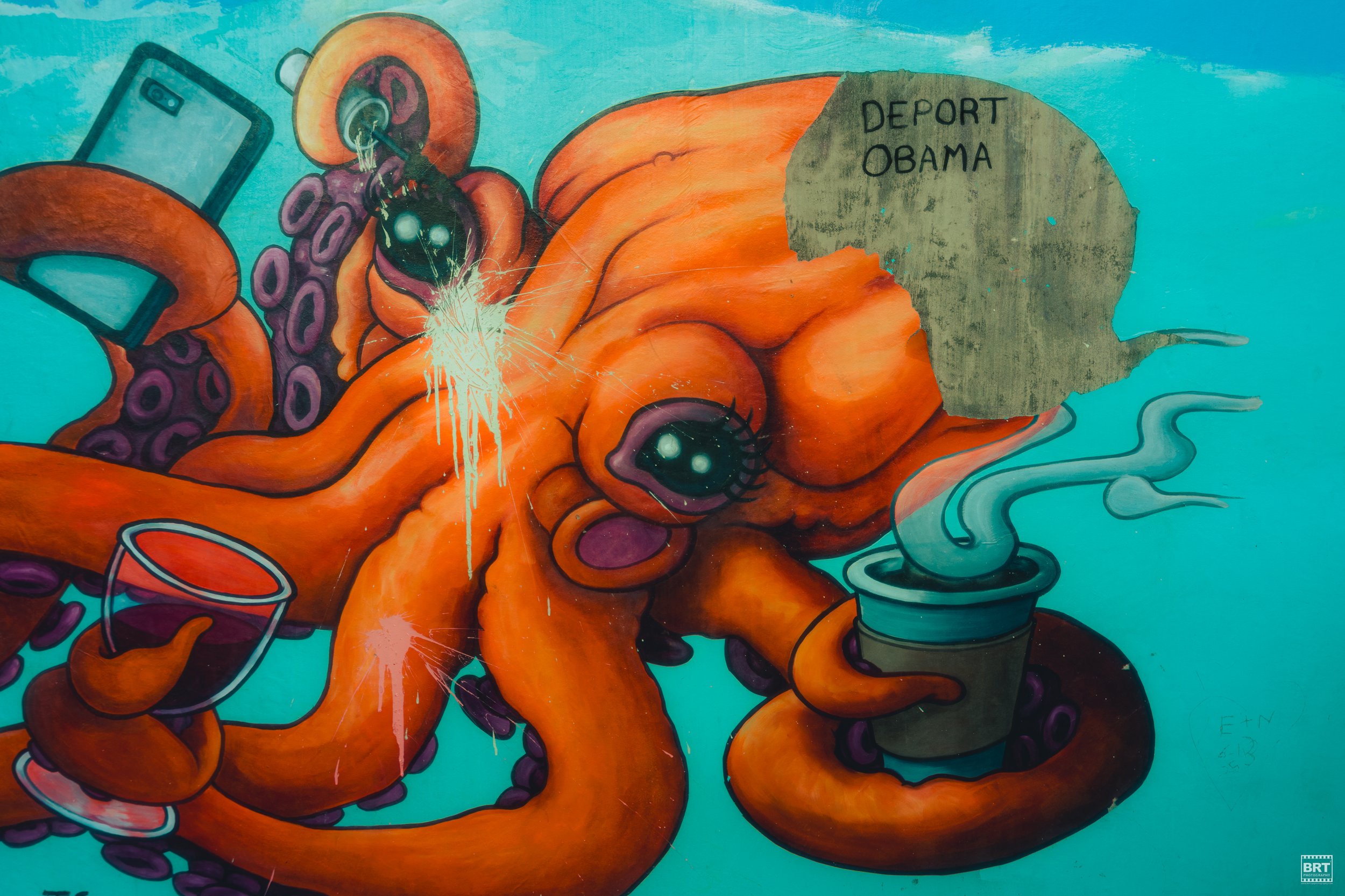 Colorful graffiti art of an orange octopus holding a smartphone and a to-go coffee cup, with purple tentacles, large cartoonish eyes, and a speech bubble with distorted text that reads 'Deport Obama' on a teal background.