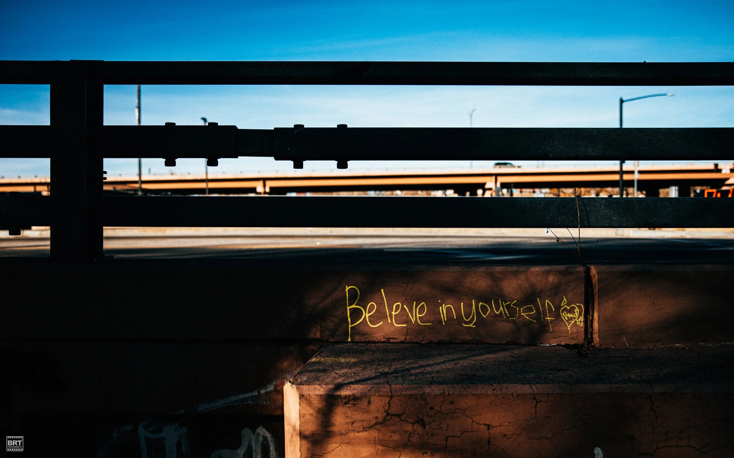 Graffiti on a concrete wall beneath a highway overpass with the message "Believe in yourself" and a small heart symbol, with the sky and highway visible in the background.