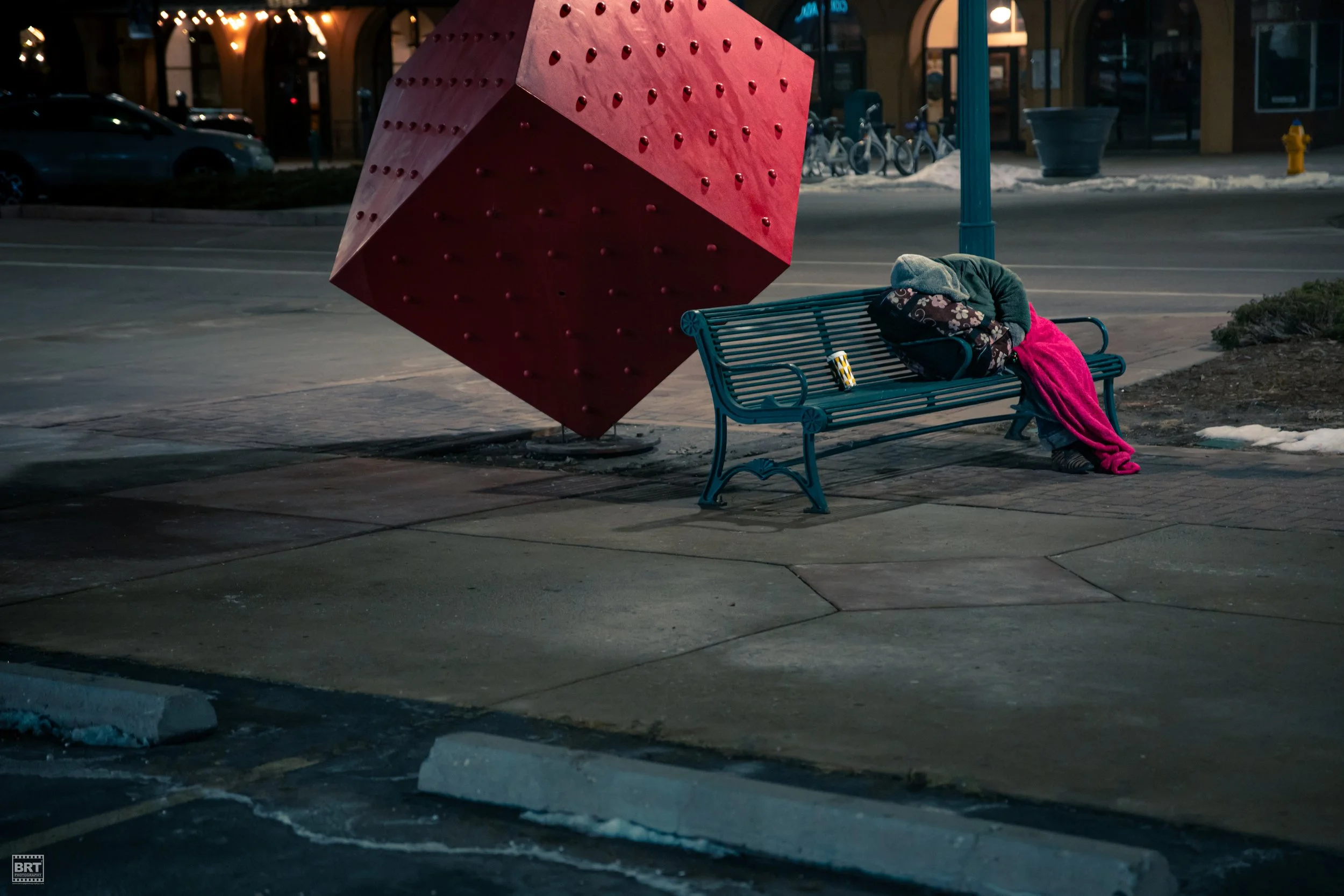 A bench with belongings, a blanket, and a backpack, is positioned beside a large red geometric sculpture on a city sidewalk at night, with cars and bicycles in the background.