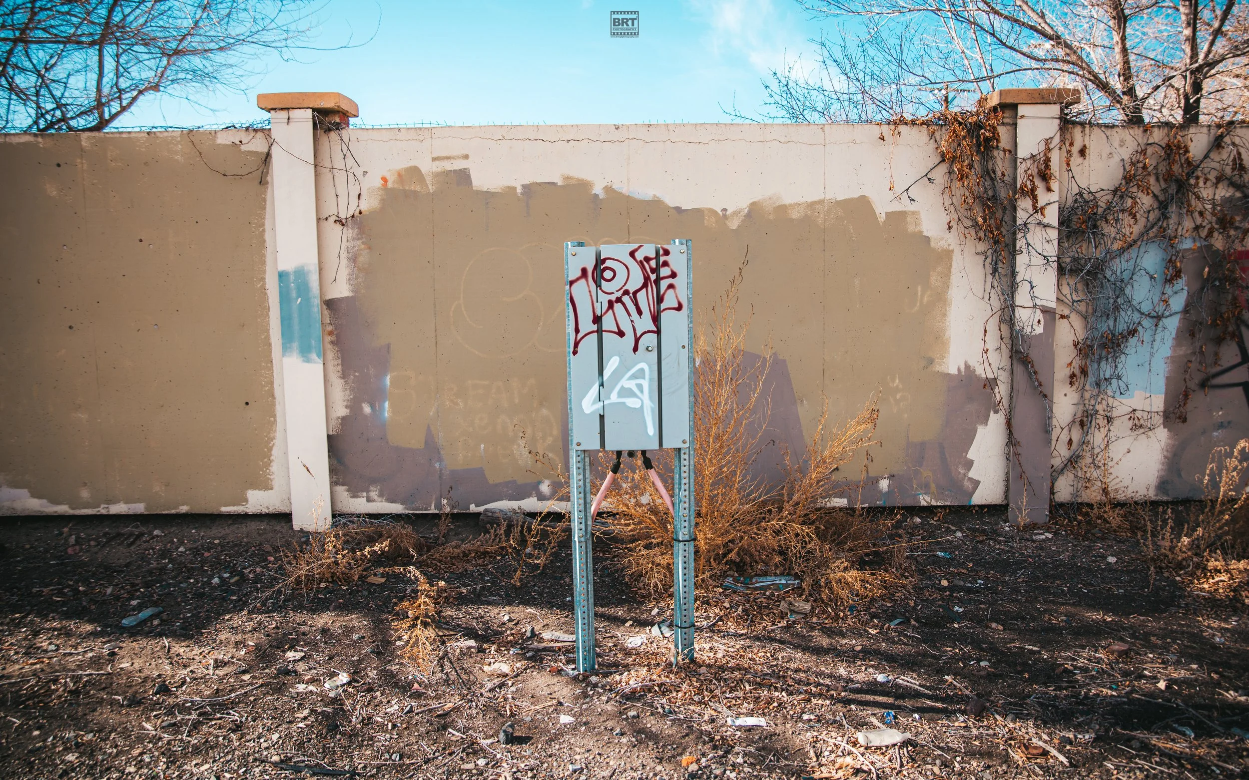 A weathered and graffiti-covered electrical panel box in front of a concrete wall with graffiti and dried bushes, with a clear blue sky above.