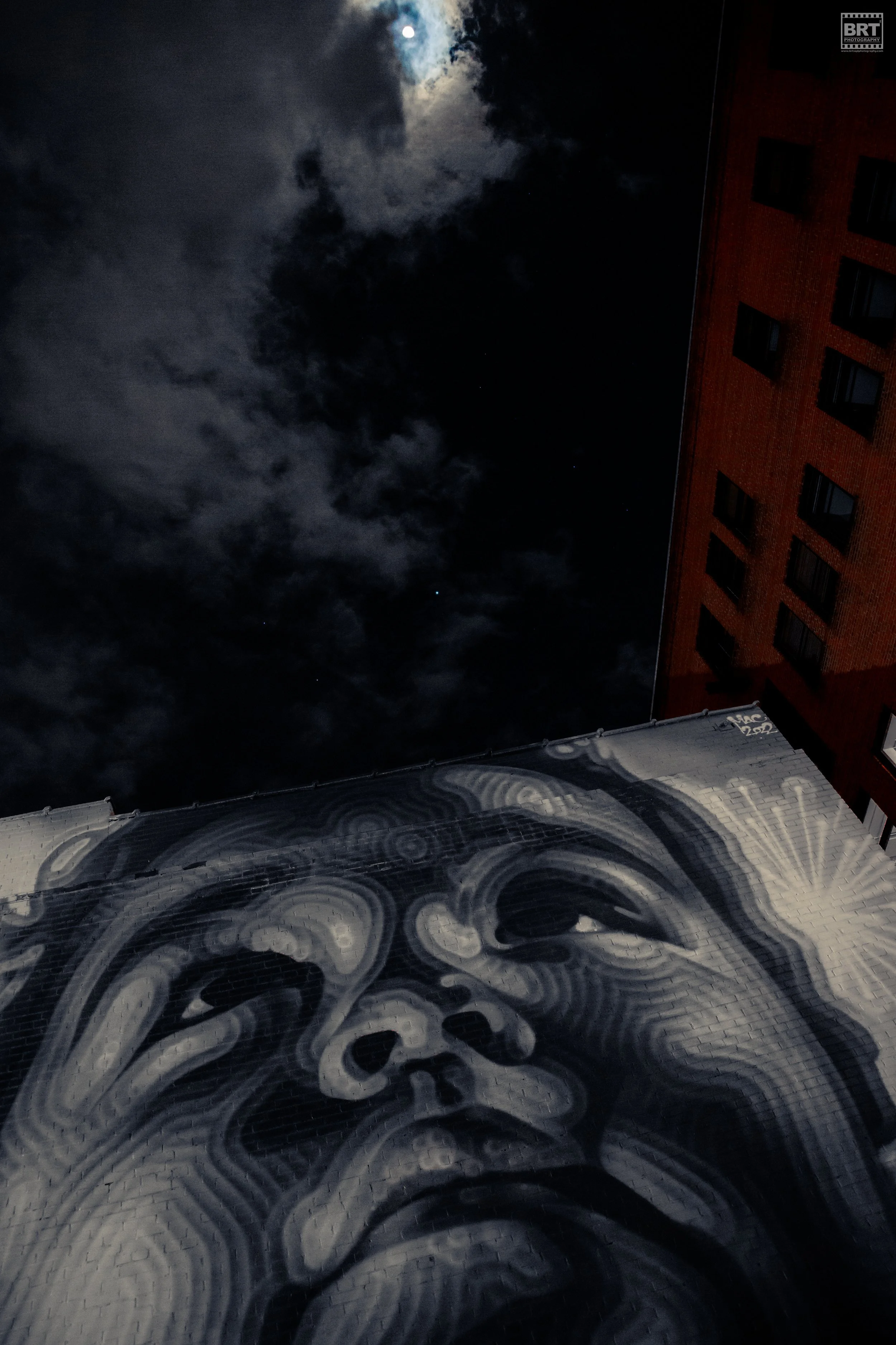 Nighttime mural of a contemplative face painted on a brick wall, with dark clouds and the moon in the night sky above.
