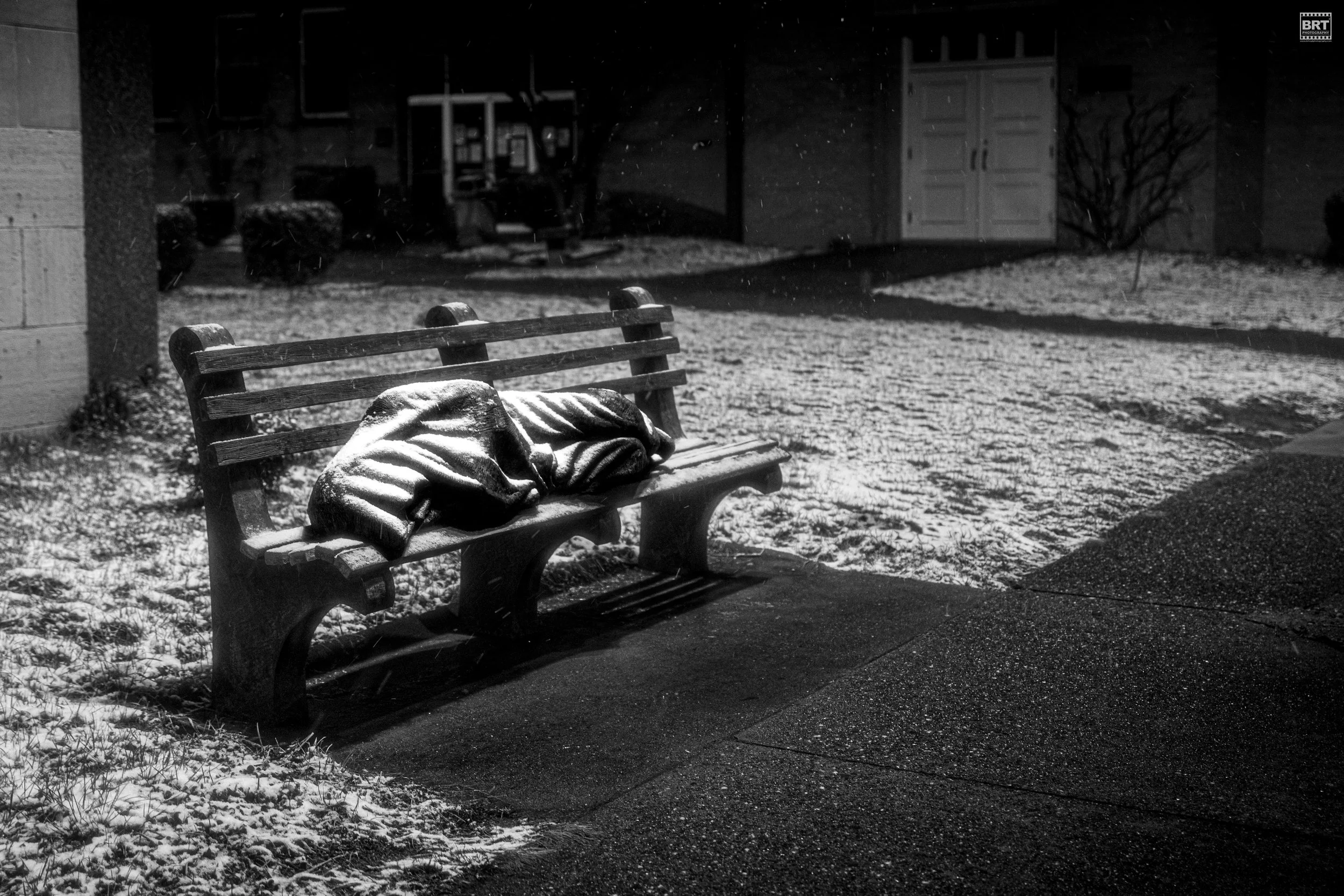 A wet park bench with a jacket on it, surrounded by snow and ice, at night.