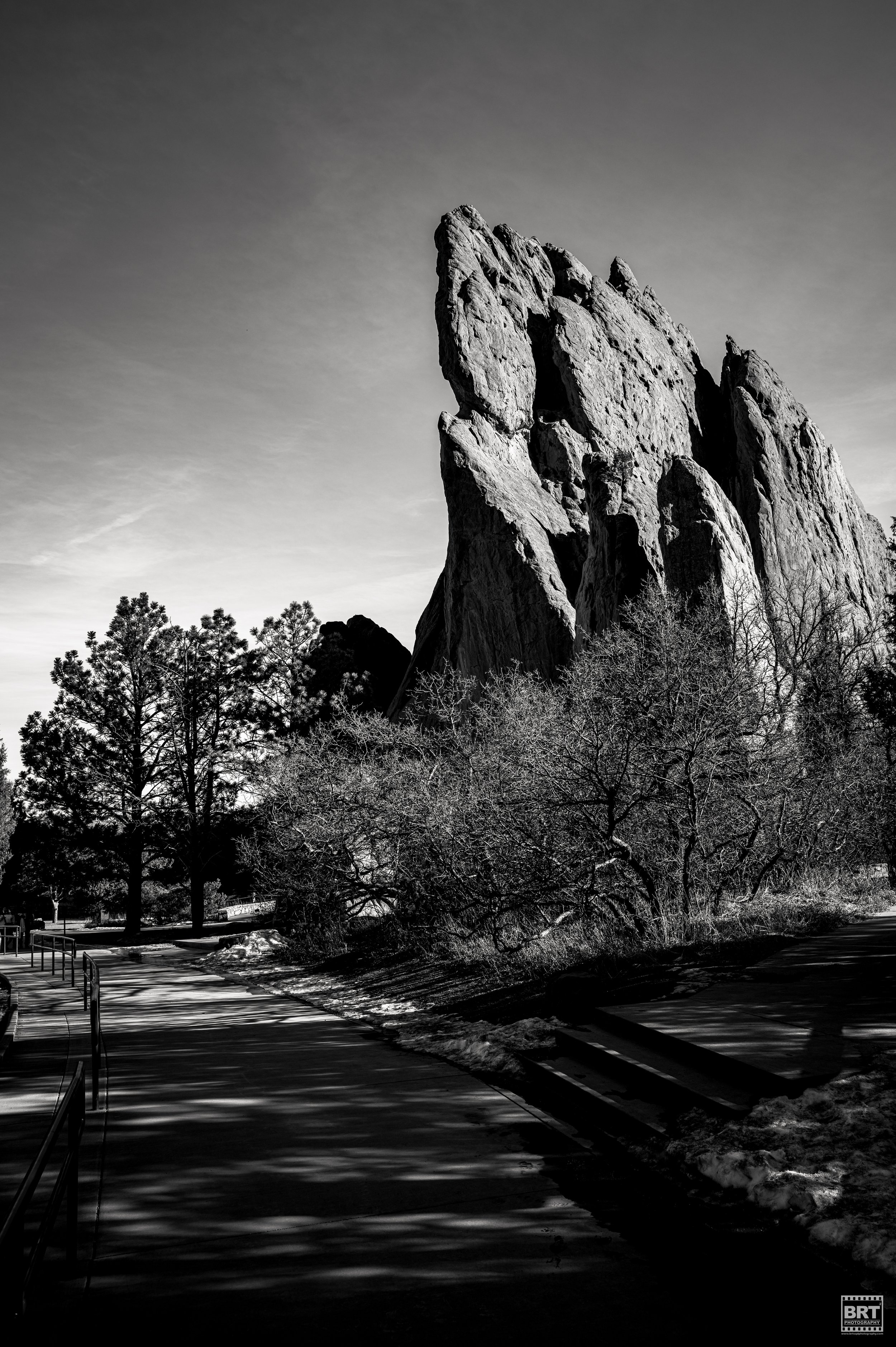 Black and white photo of a large rock formation with trees in the foreground and a paved pathway.